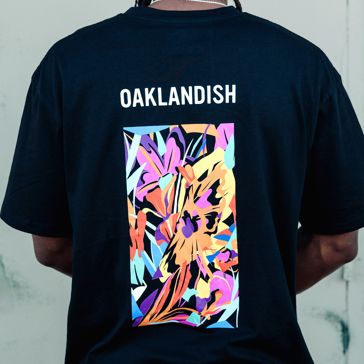 A person viewed from behind wears a black Urban Tropics Tee, featuring an abstract design of colorful tropical leaves and the word “OAKLANDISH” in bold white text against a plain, light-colored background.