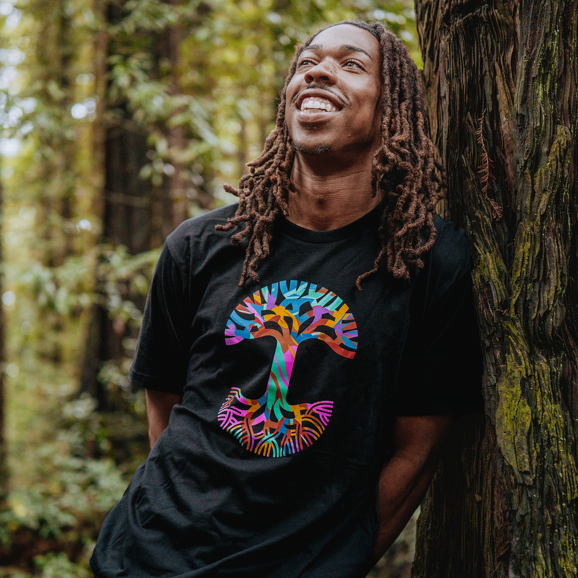 A smiling man with long dreadlocks leans against a tree in a lush forest, wearing the Oaklandish Urban Tropics Tee—an abstract art t-shirt with a large, vibrant mushroom graphic in blue, pink, orange, and green.