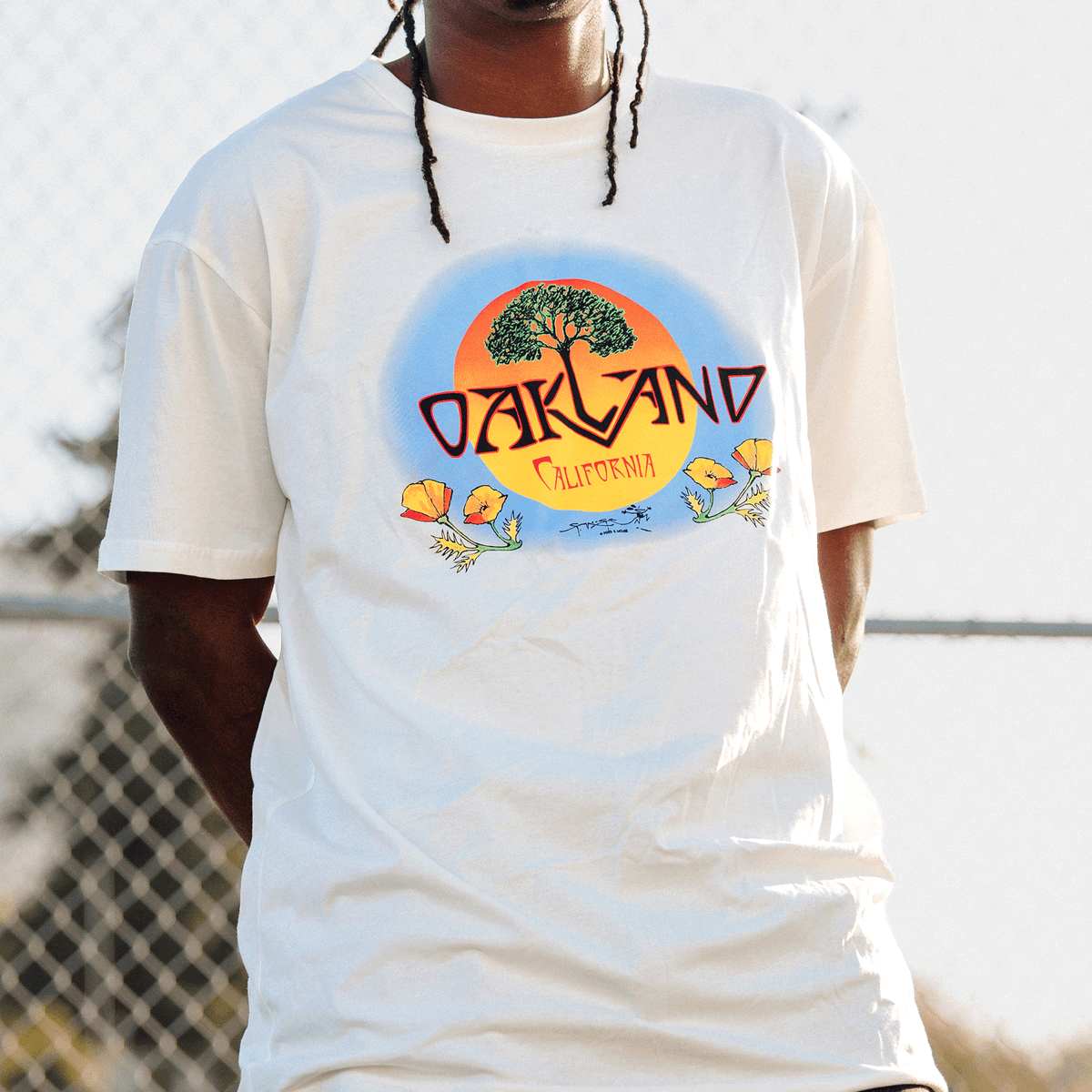 A person wears a cream-colored Oaklandish Vanguard Tee featuring a chest graphic of a green tree against a yellow and blue sun, with “OAKLAND” in bold orange, “CALIFORNIA” below it, and stylized orange poppies at the bottom.