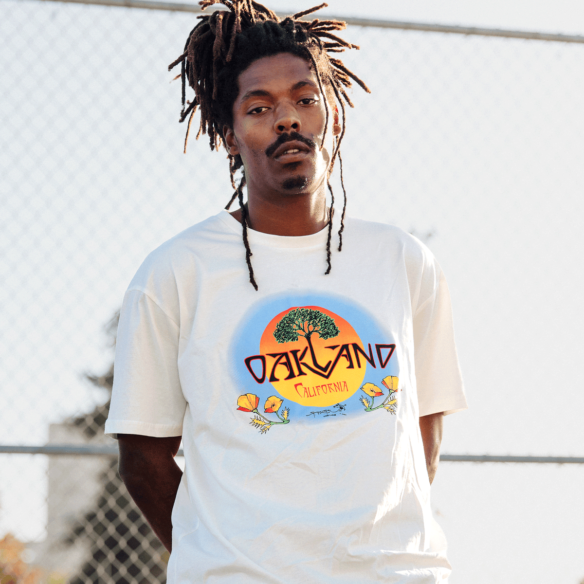 A man with dreadlocks wears the Oaklandish Vanguard Tee, a white t-shirt featuring a graphic of a sun setting behind a tree with “OAKLAND CALIFORNIA” and orange flowers, standing outdoors in front of a chain-link fence and looking at the camera.