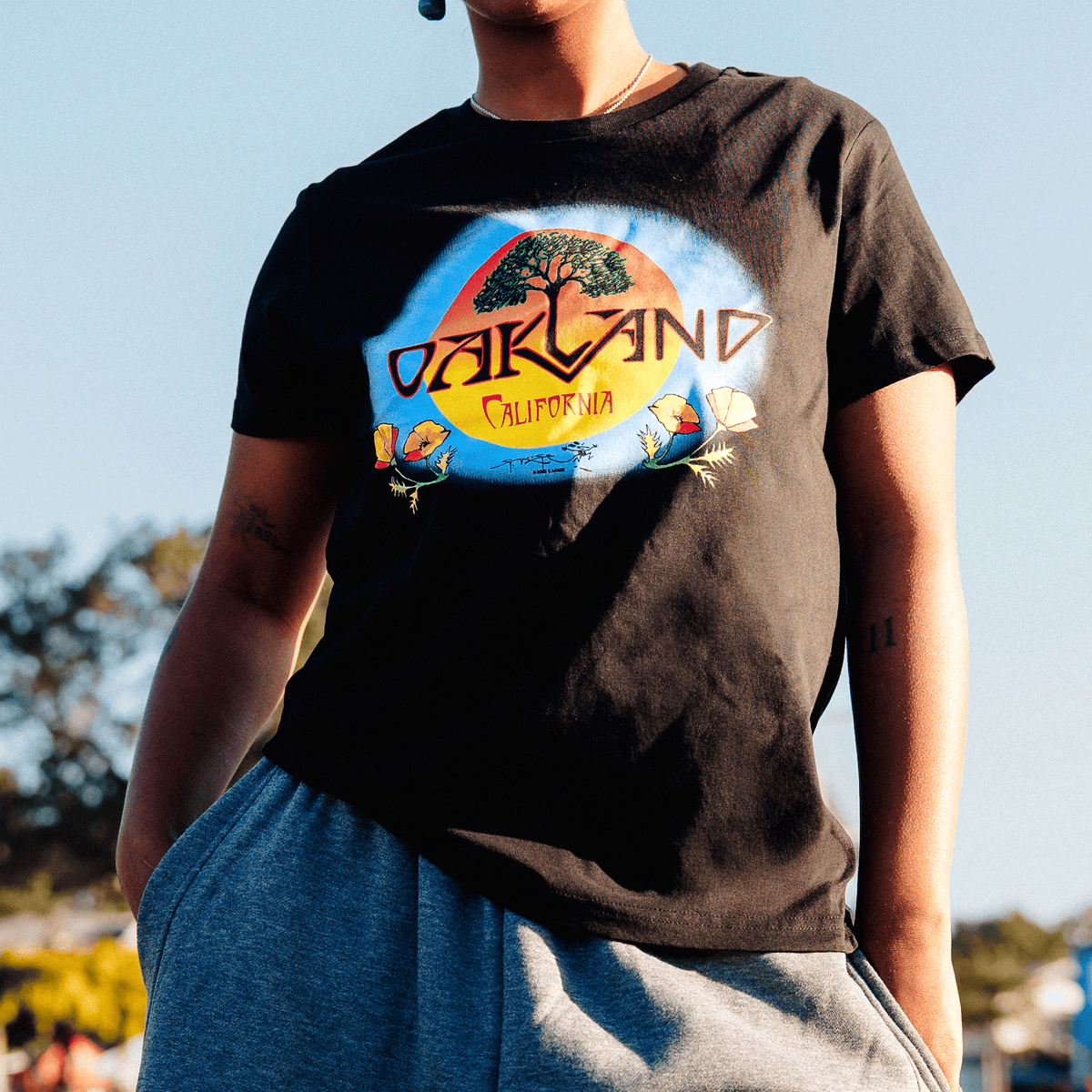 A person wears a black Women’s Vanguard Tee by Oaklandish, featuring a Stanley Mouse graphic of an oak tree inside a sun-like circle, with “OAKLAND CALIFORNIA” text and poppy illustrations set against a hazy sky.