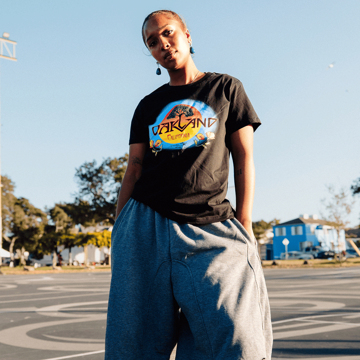 A person with braided hair wears a black Women’s Vanguard Tee by Oaklandish, featuring a circular tree silhouette against a sun with “Oakland” arched above, standing in a paved lot during golden hour in grey sweatpants with hands in their pockets.