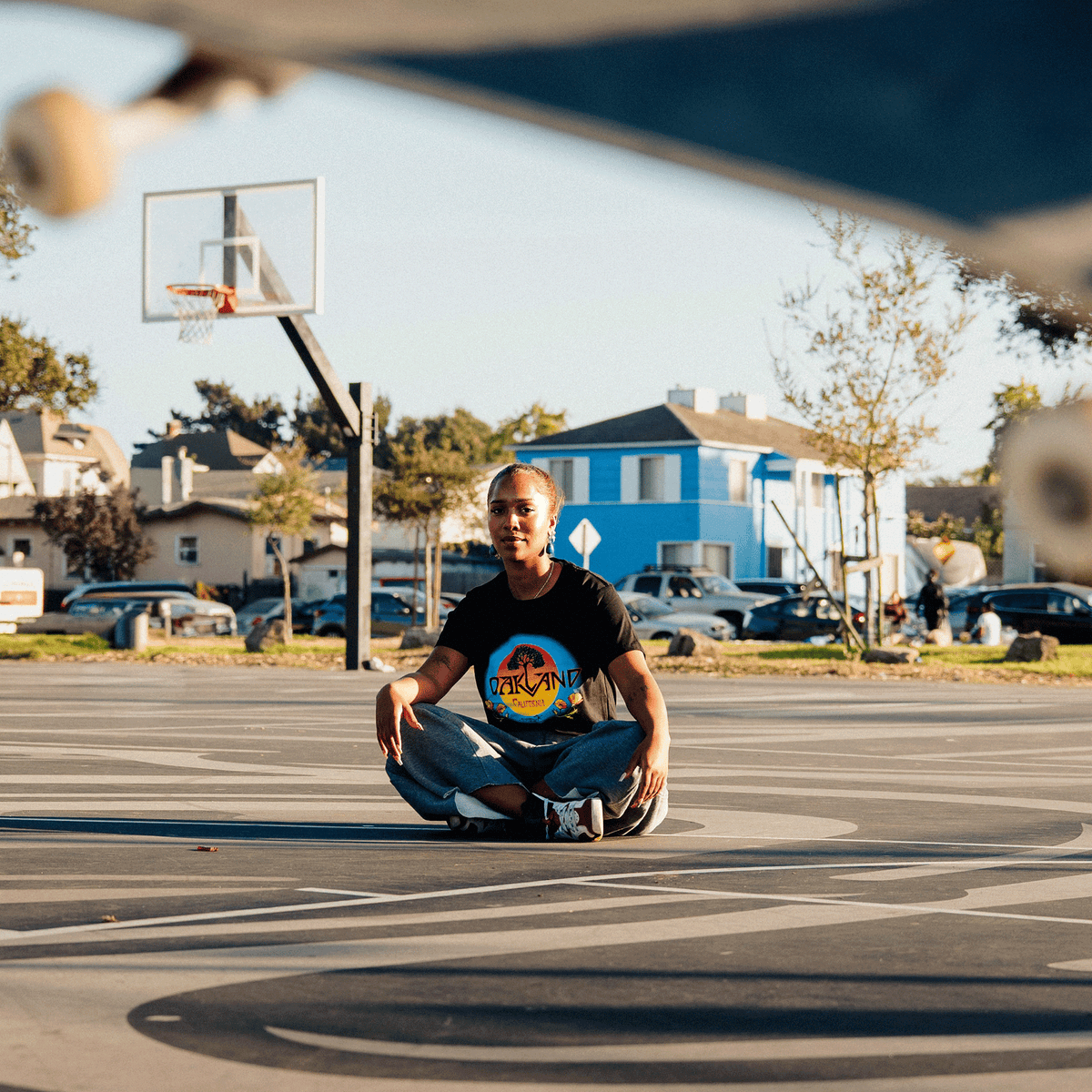 A person with braided hair wears a black Oaklandish Women’s Vanguard Tee and loose-fitting jeans while sitting cross-legged on a basketball court, centered with a hoop and suburban houses in the background, partially framed by a skateboard deck.