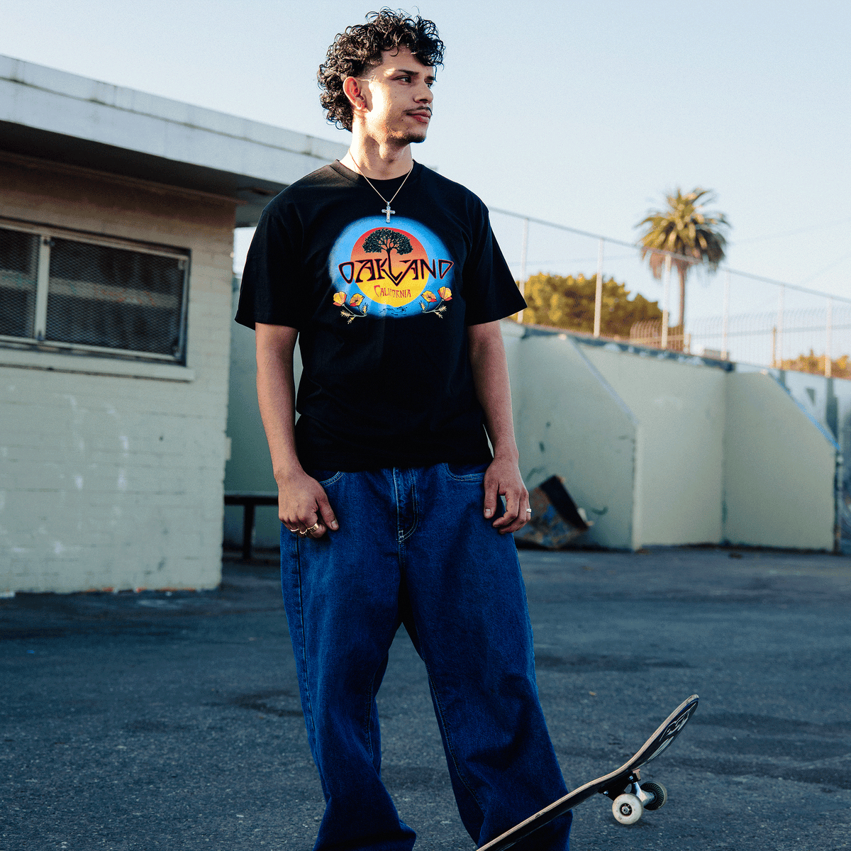 A young man with curly dark hair stands outdoors on a paved lot with a skateboard at his feet, wearing an Oaklandish Vanguard Tee featuring a tree inside a circular sunset design, loose-fit blue jeans, and a silver cross necklace near a tan building and fence.