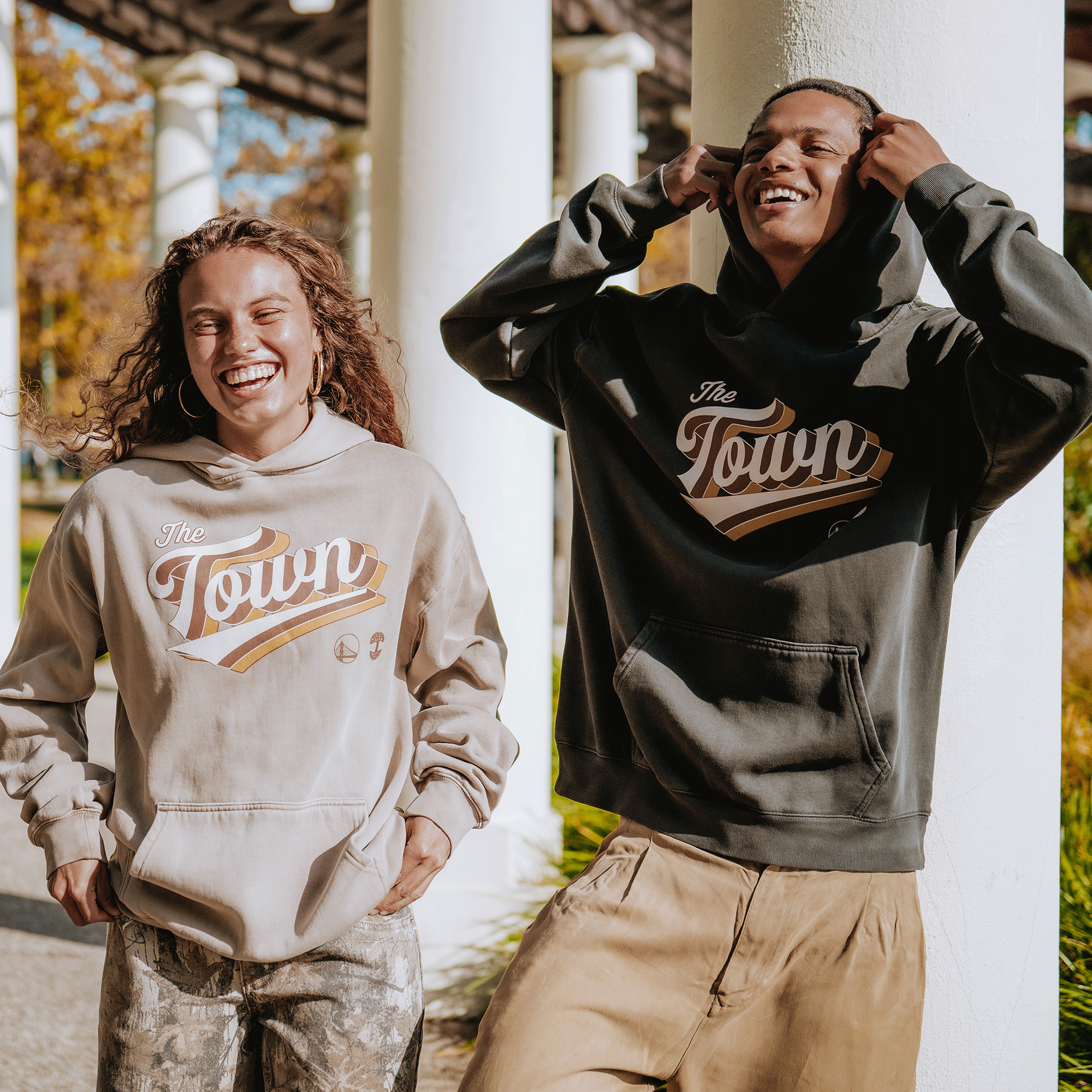 Two people stand outdoors, smiling in cozy GSW Vintage Swash Hoodies—one in light beige with patterned pants, the other in olive green with tan pants. Retro graphics pop on both hoodies. White columns are visible behind them.