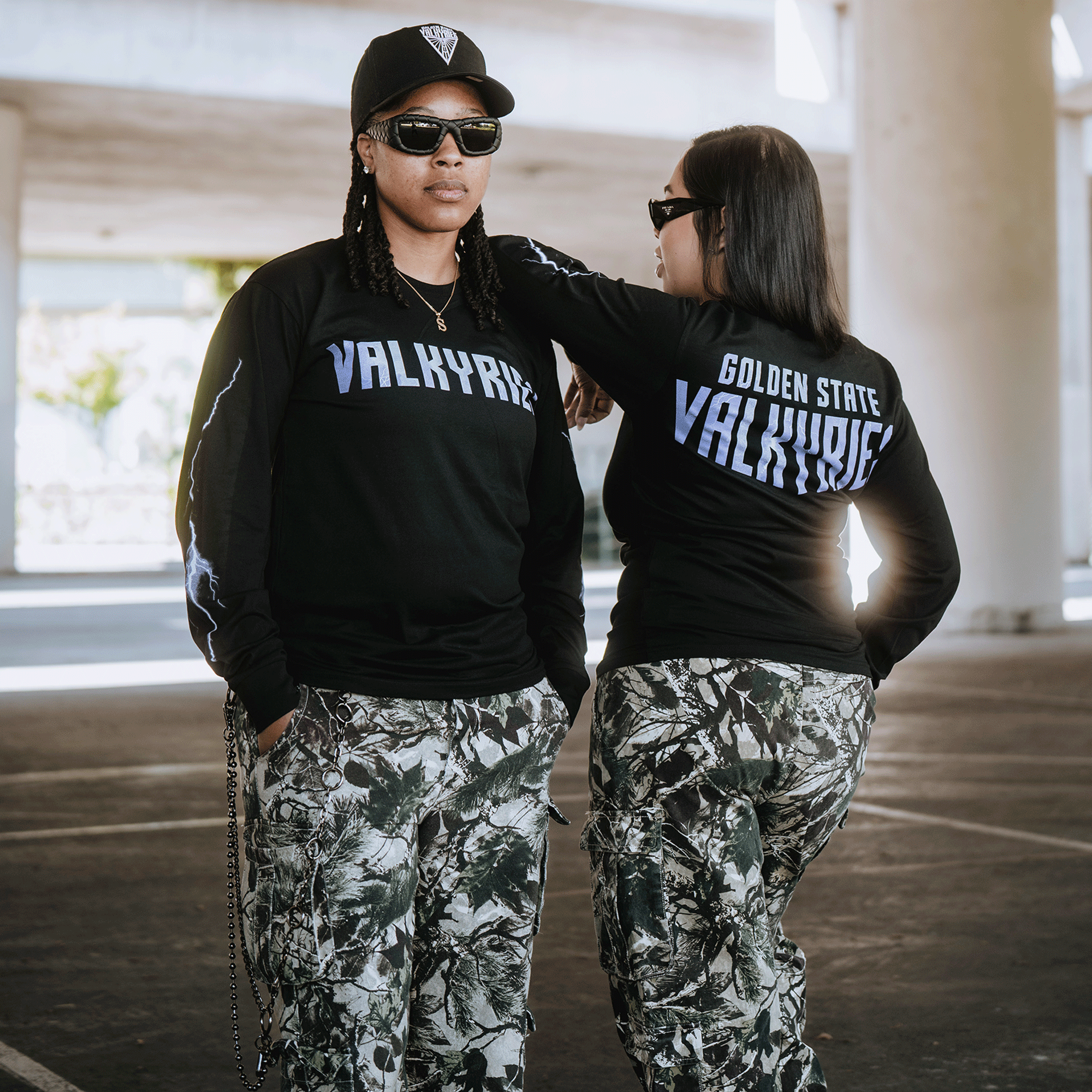 Two people pose confidently in the city, both wearing black GSV Golden State Valkyries Violet Lightning LS Tees and camouflage pants. The back of one shirt features "Golden State Valkyries" as they face away from the camera.
