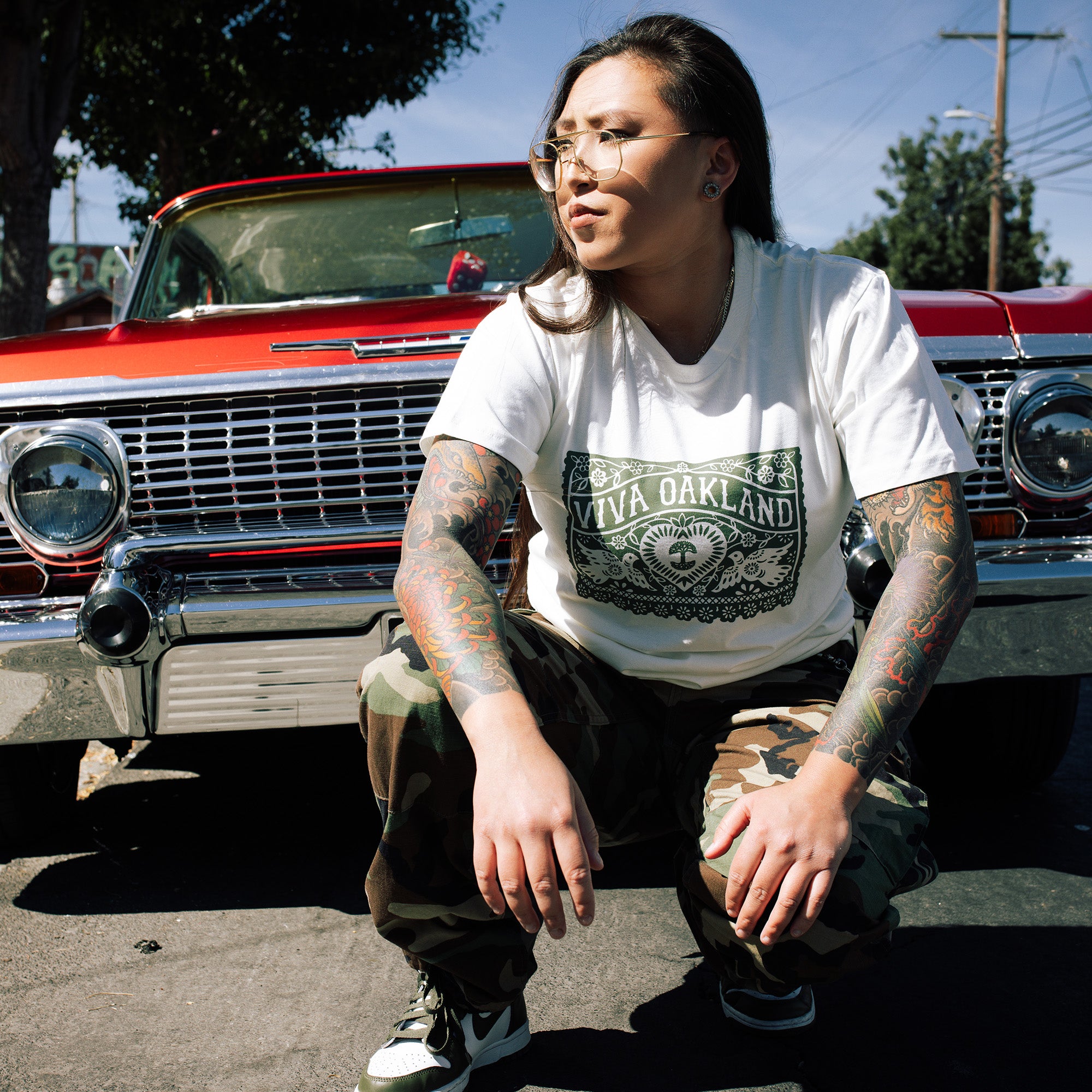 A person with long hair, glasses, and colorful arm tattoos squats in front of a vintage red car. They are wearing an Oaklandish "Viva Oakland Tee" made from 100% cotton, paired with camouflage pants and black-and-white sneakers. The background showcases a tree, some greenery, and a clear sky.