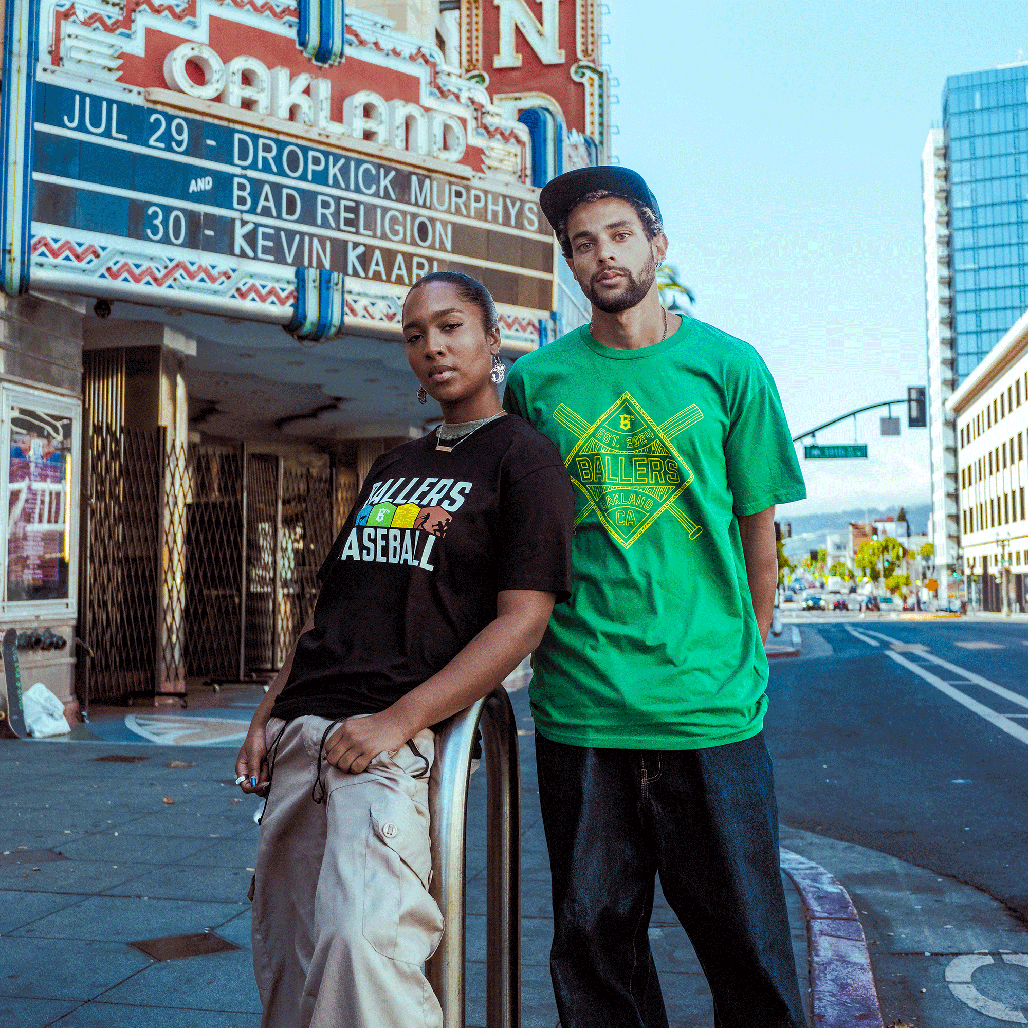 Two people stand on a city sidewalk in front of an Oakland theater marquee. The woman wears the Oakland Ballers Classic Diamond Tee; the man has on a green shirt and blue cap. Street life and tall buildings complete the scene.