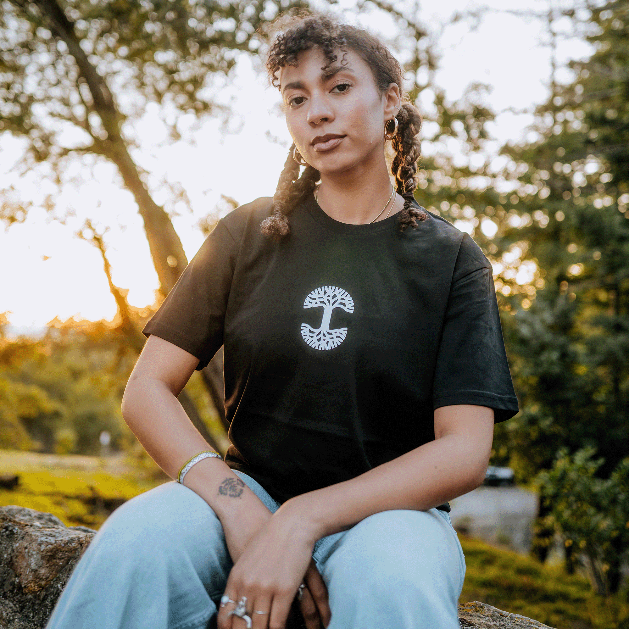 As the sun sets, casting a warm glow on the trees and sky similar to Oakland's charm, a person with curly hair sits outdoors. They wear an Oaklandish Who We Are and Who We Do it For Tee, which hugs their silhouette in a classic fit, showcasing a circular decorative logo in its center. Hoop earrings glint in their ears while bracelets and rings adorn their arms resting on their knees.