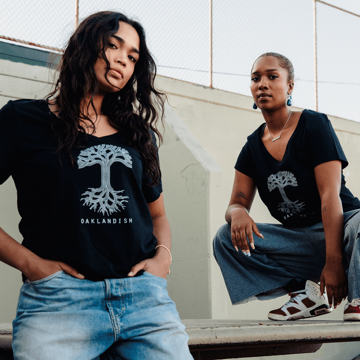 Two women wear Women’s Oaklandish Logo V-Neck t-shirts, which are 100% cotton and feature a white graphic of an oak tree with exposed roots. They pose against a light-colored concrete wall and chain-link fence, styled with casual jeans and sneakers.