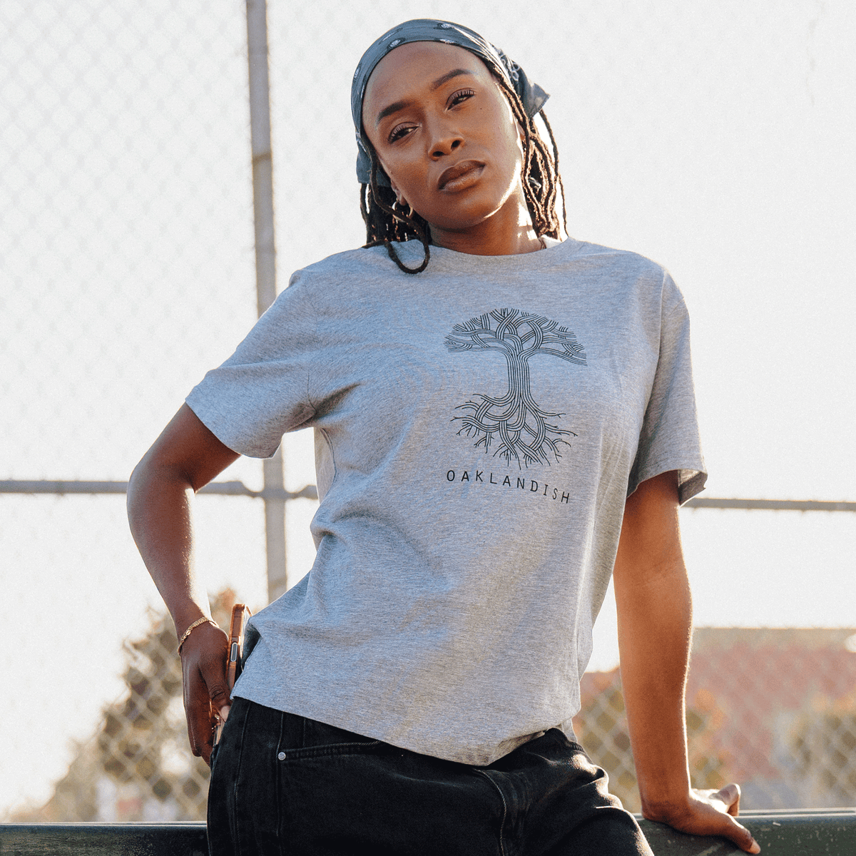 A woman with long dark braids and a grey bandana leans against an outdoor fence, wearing a Women’s Oaklandish Logo Tee in heather grey with dark jeans.
