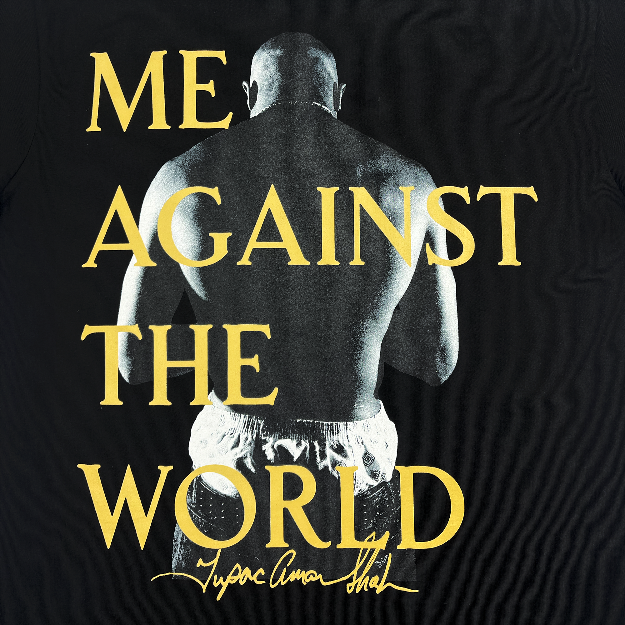 The Against the World Tee by Oaklandish is a black T-shirt inspired by hip-hop culture, featuring a grayscale image of a muscular figure from behind. Bold yellow letters display "ME AGAINST THE WORLD" across the design, with Tyson Fury's name in signature-like script below, capturing Tupac's Oaklandish spirit.