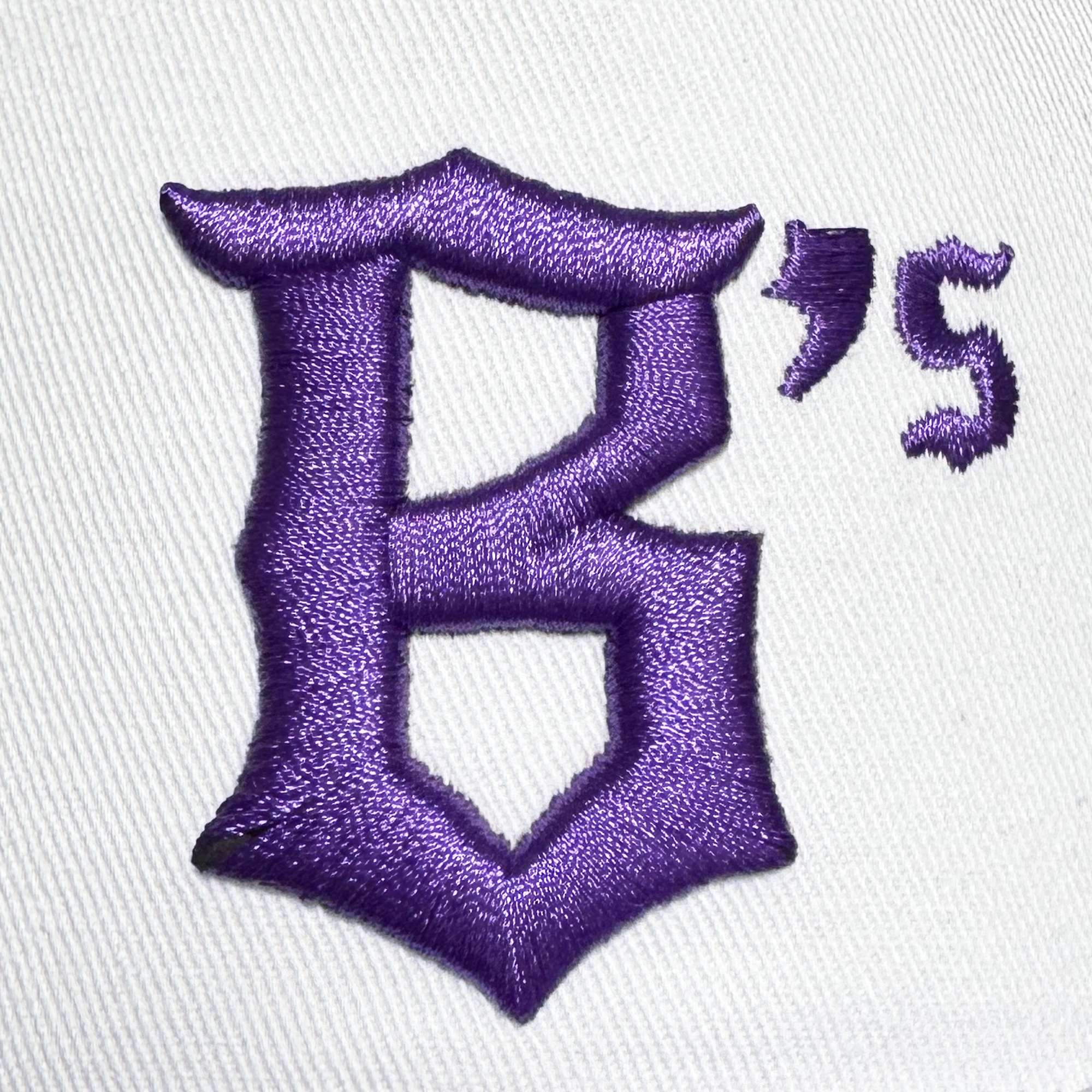 A raised, textured purple “B” in Old English script, followed by a smaller matching “’s,” is embroidered onto a white fabric background on the Oakland Ballers 5 Panel Cap by Oakland Ballers. The design echoes the iconic style of the team’s signature snapback hat.