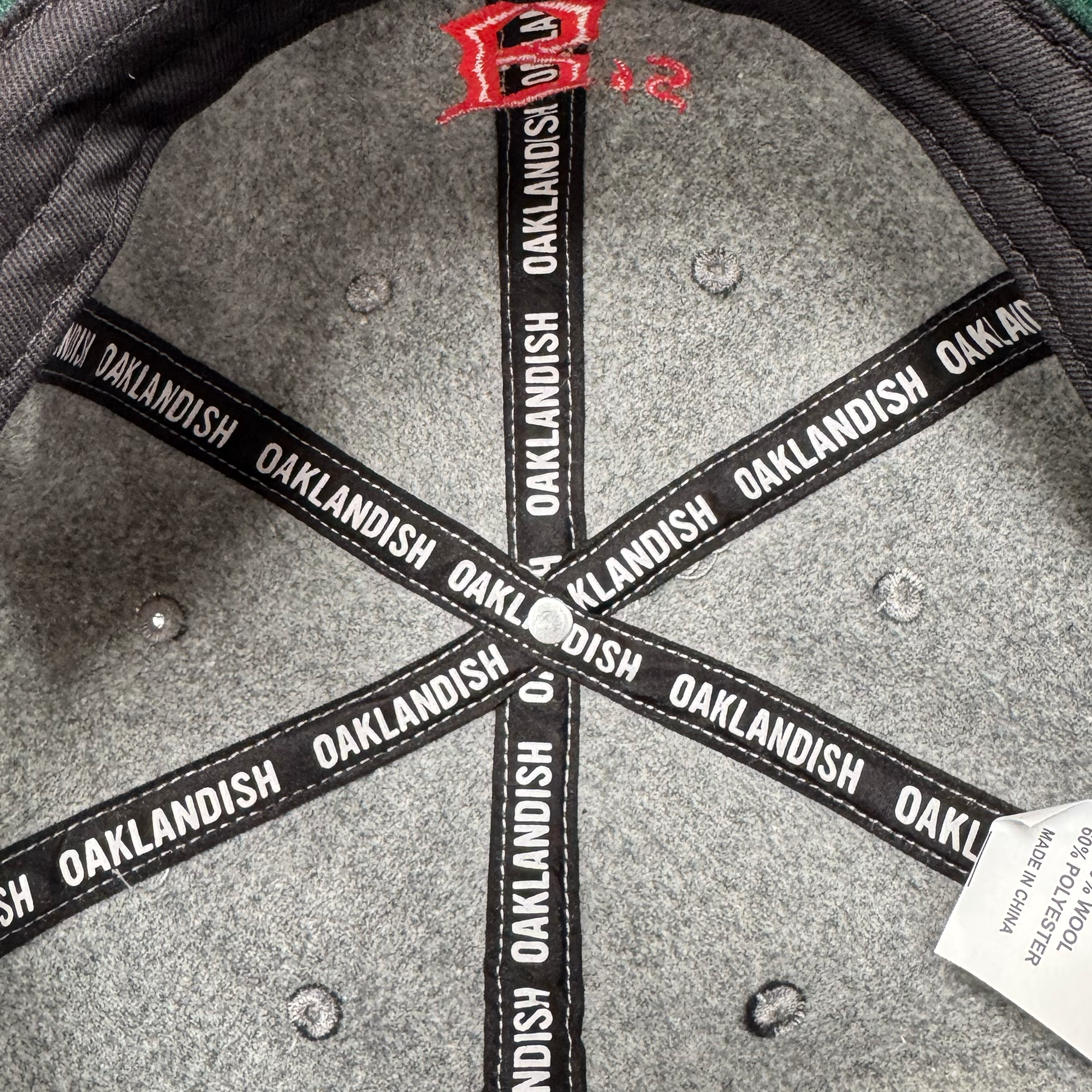 The interior of the Oakland Ballers B Strapback by Oakland Ballers features gray polyester wool, black hat bands with repeated white "OAKLANDISH" text, six silver rivets, a right-side white tag, and red embroidered stitching in the crown.