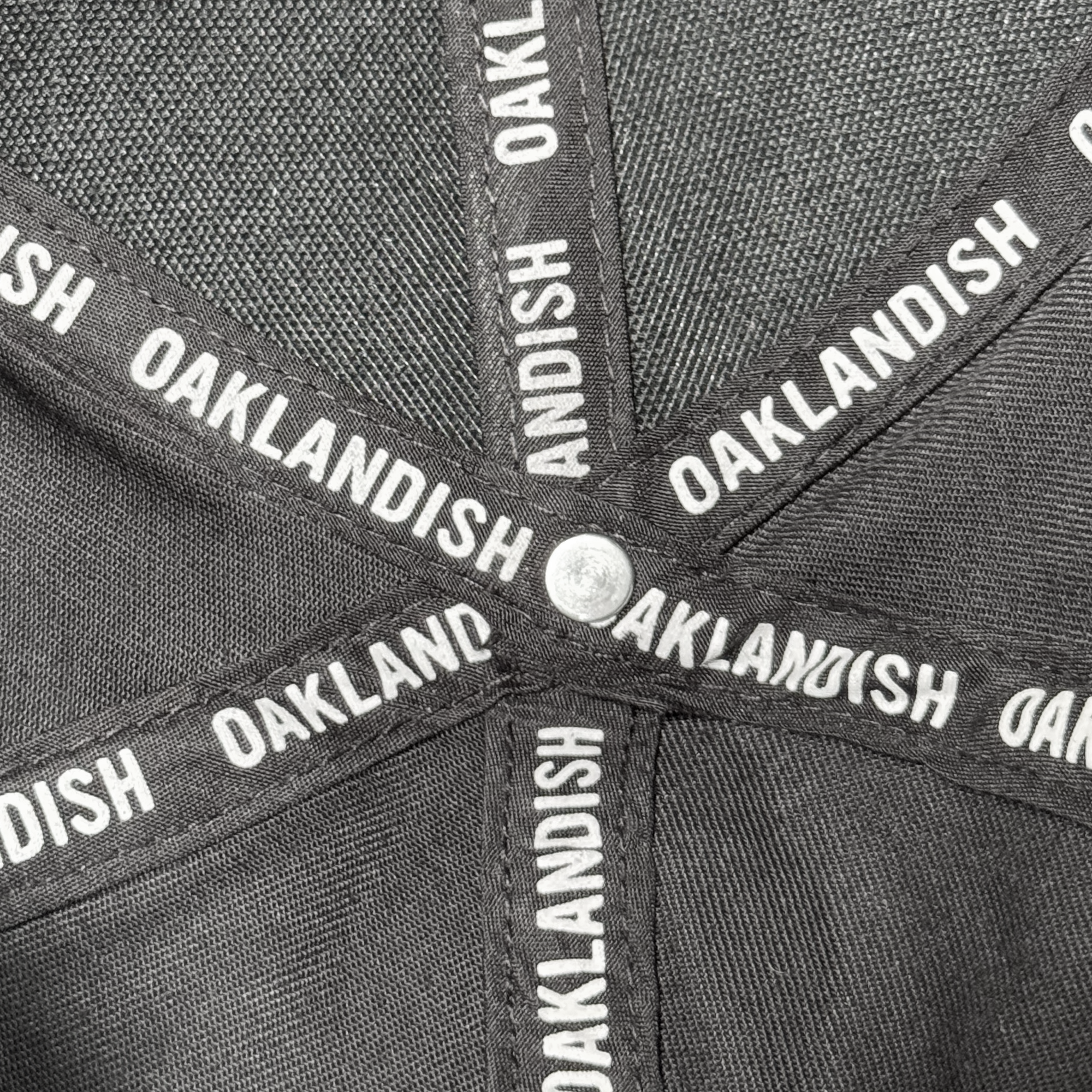 Close-up of the inside of an Oakland Ballers Flexfit Cap, showing intersecting black fabric bands with "OAKLANDISH" in white, a small silver center button, and mesh polyester fabric in the background. Brand: Oakland Ballers.