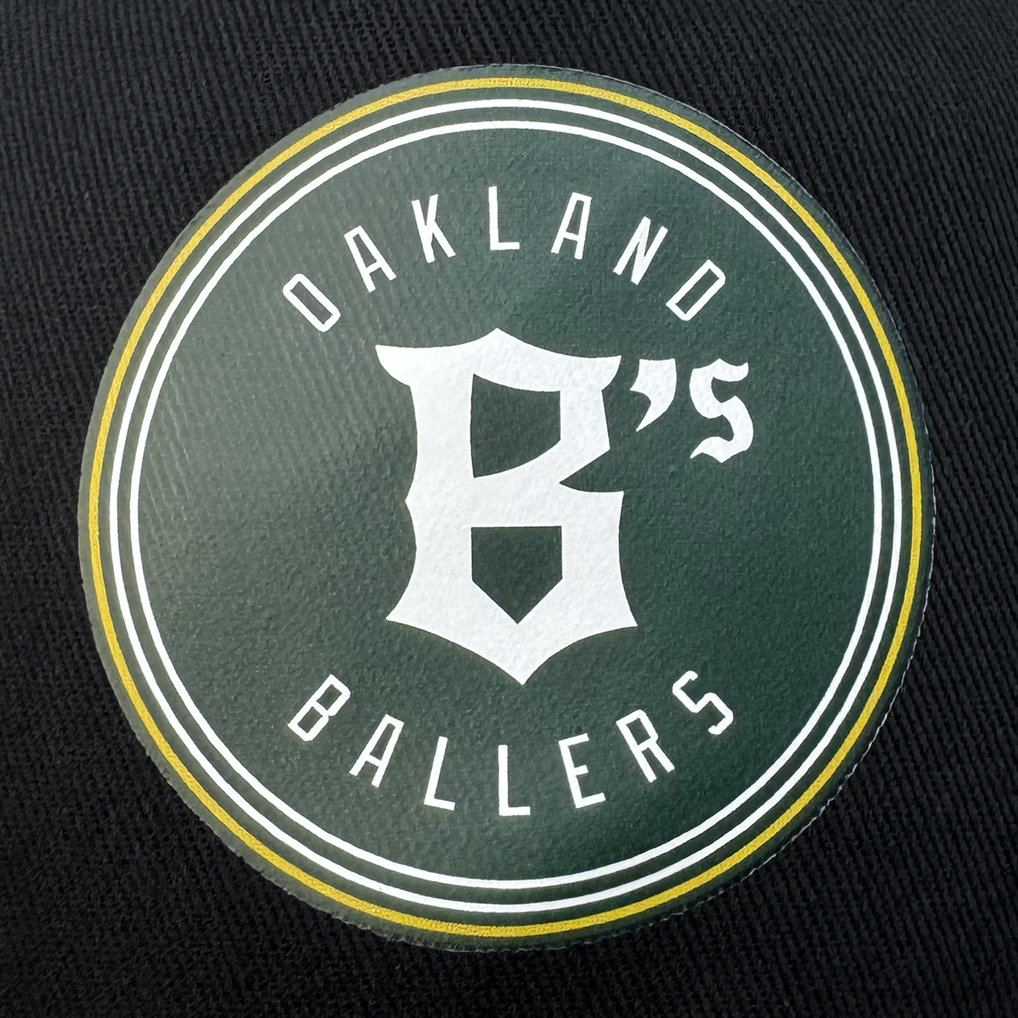 The Oakland Ballers Classic Golf Cap features a dark green emblem with a large white "B" on a black background, bordered by white and encircled in yellow. The text "OAKLAND BALLERS" represents Oakland's new professional baseball team in the Pioneer League.