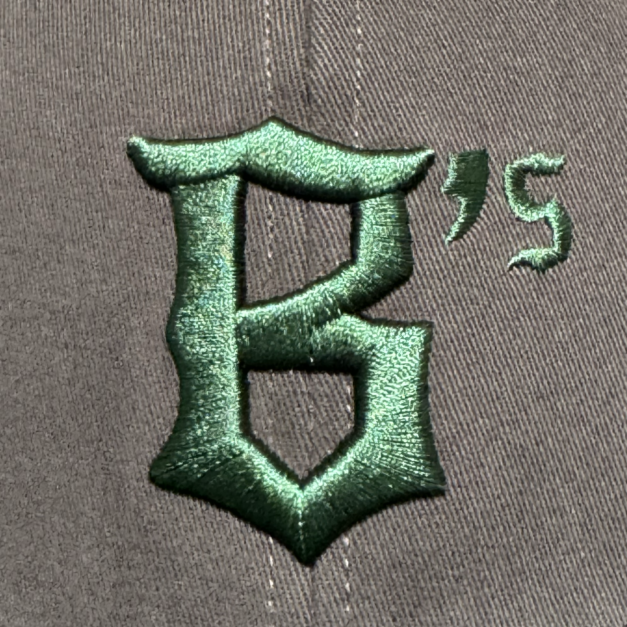 A close-up of the Oakland Ballers Logo Hat by Oakland Ballers shows gray fabric with a large dark green embroidered gothic "B," plus an apostrophe and "S," representing the new Oakland Pioneer League baseball team’s logo.