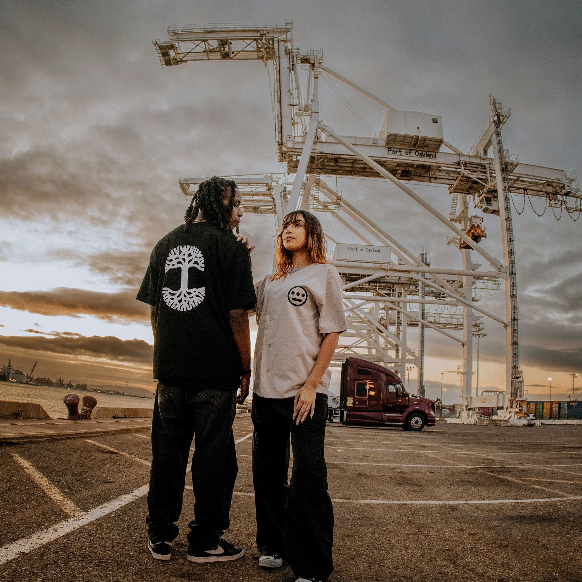 Male model's backside view of black baseball jersey with large white Oaklandish tree logo print in the center standing with female model in hiero baseball jersey.