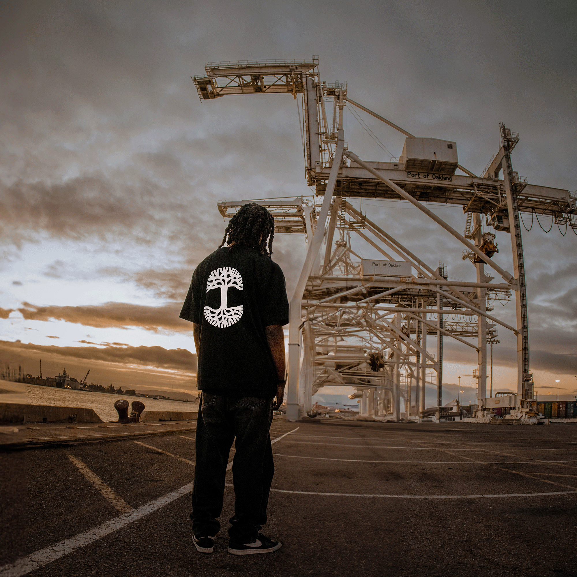A person with long hair stands facing away, wearing an Oaklandish Heavy Button-up Baseball Jersey, dark pants, and sneakers. They are in an industrial port area during sunset, with large cranes and machinery visible in the background under a cloudy sky.