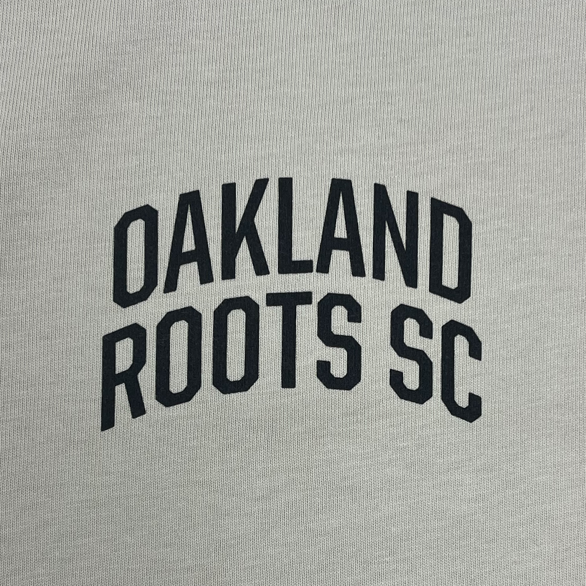 Close-up of black OAKLAND ROOTS SC wordmark on the left chest of a bone-colored button-up baseball jersey. 