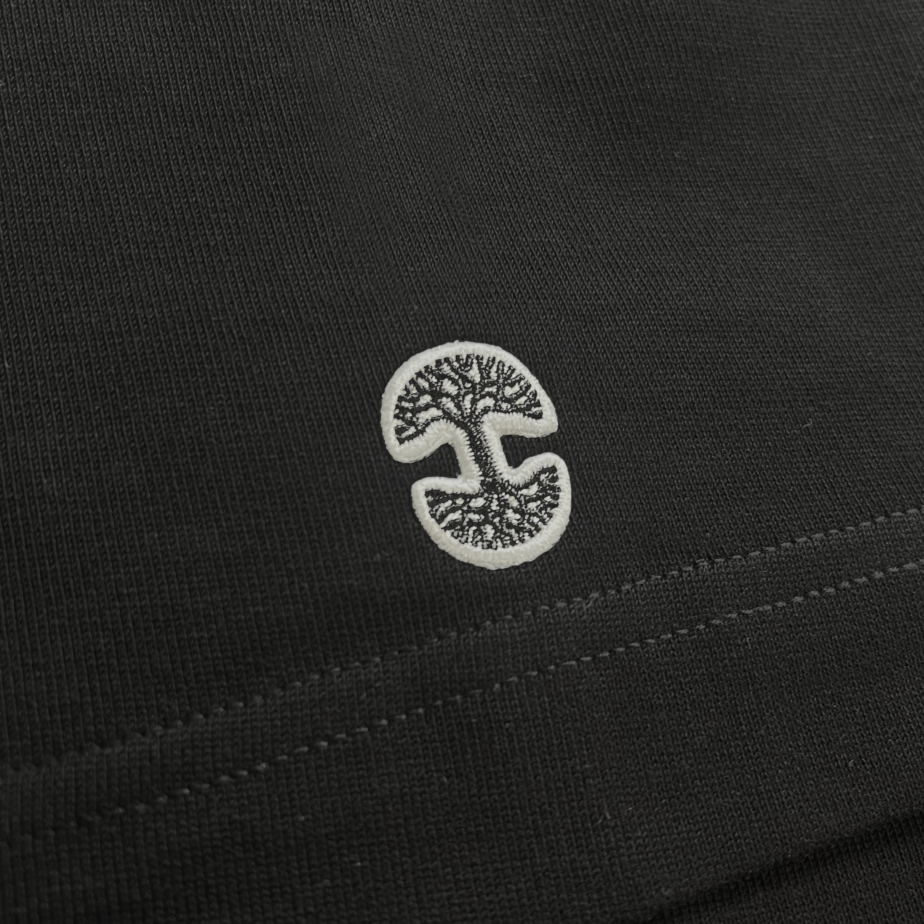 A close-up view of the black fabric surface on the Oaklandish Heavy Button-up Baseball Jersey, showcasing the Oaklandish tree logo. The logo features two mirrored tree-like patterns facing each other, creating an abstract, symmetrical design. The detailed stitching and texture of the fabric are clearly visible, indicating it's men’s sizing from Oaklandish.