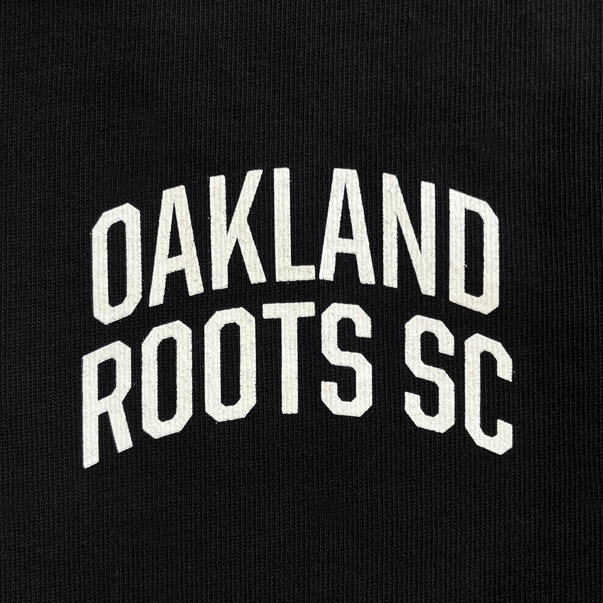 Detailed close-up of white OAKLAND ROOTS SC wordmark on a black baseball jersey.