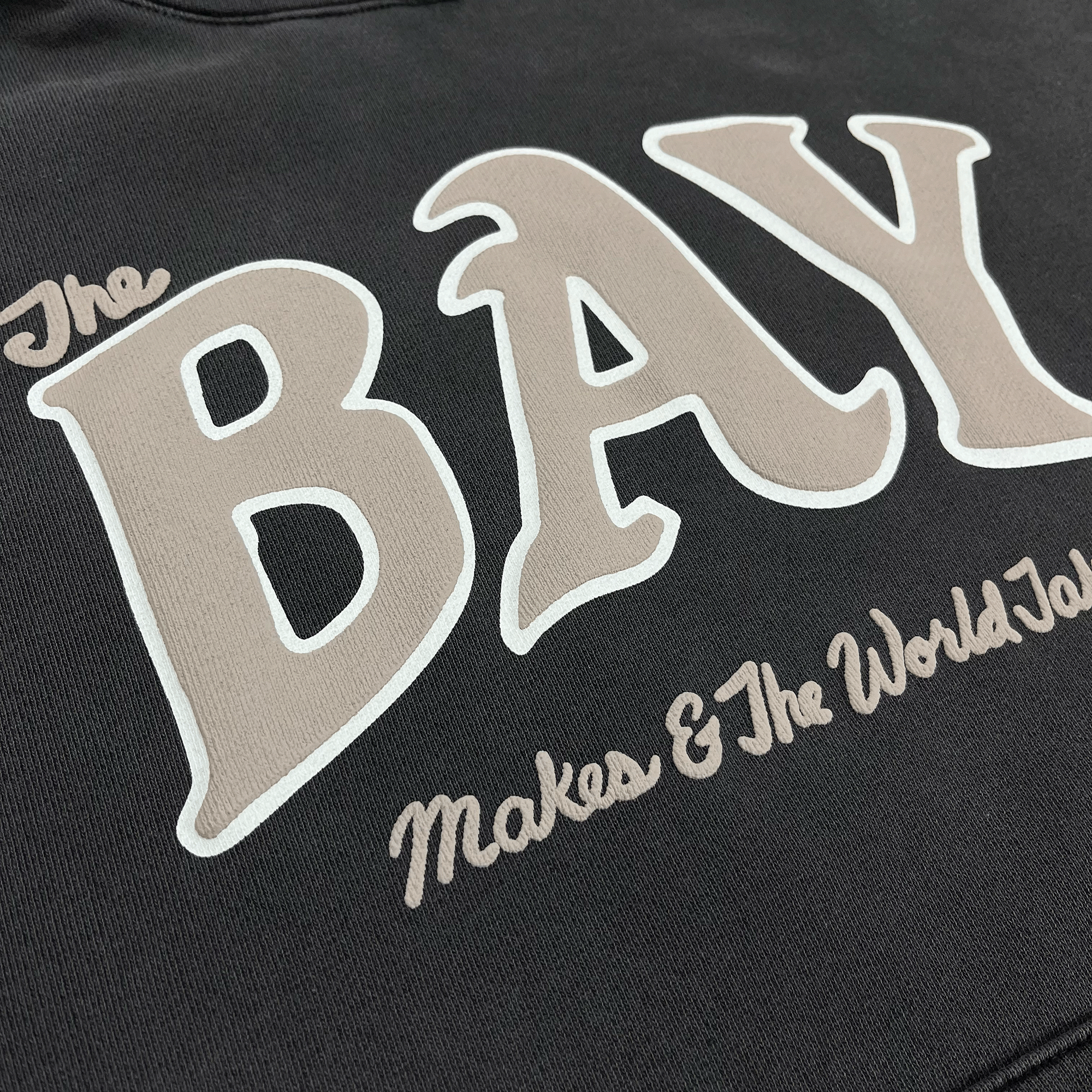 Close-up of tan and white The Bay Makes, The World Takes logo on the front chest of a pigment black hooded sweatshirt.