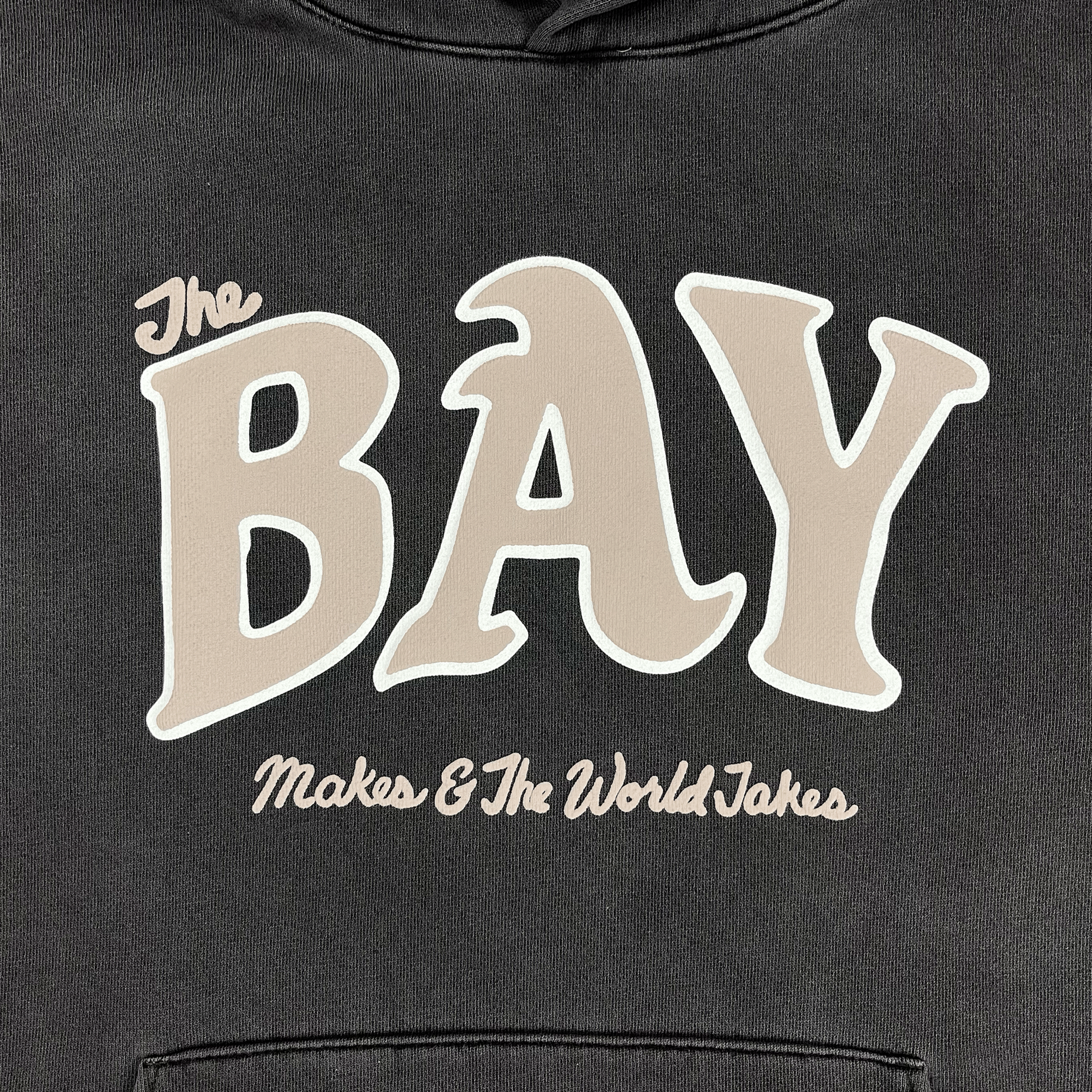  Close-up of tan and white The Bay Makes, The World Takes logo on the front chest of a  pigment black pullover hoodie sweatshirt.