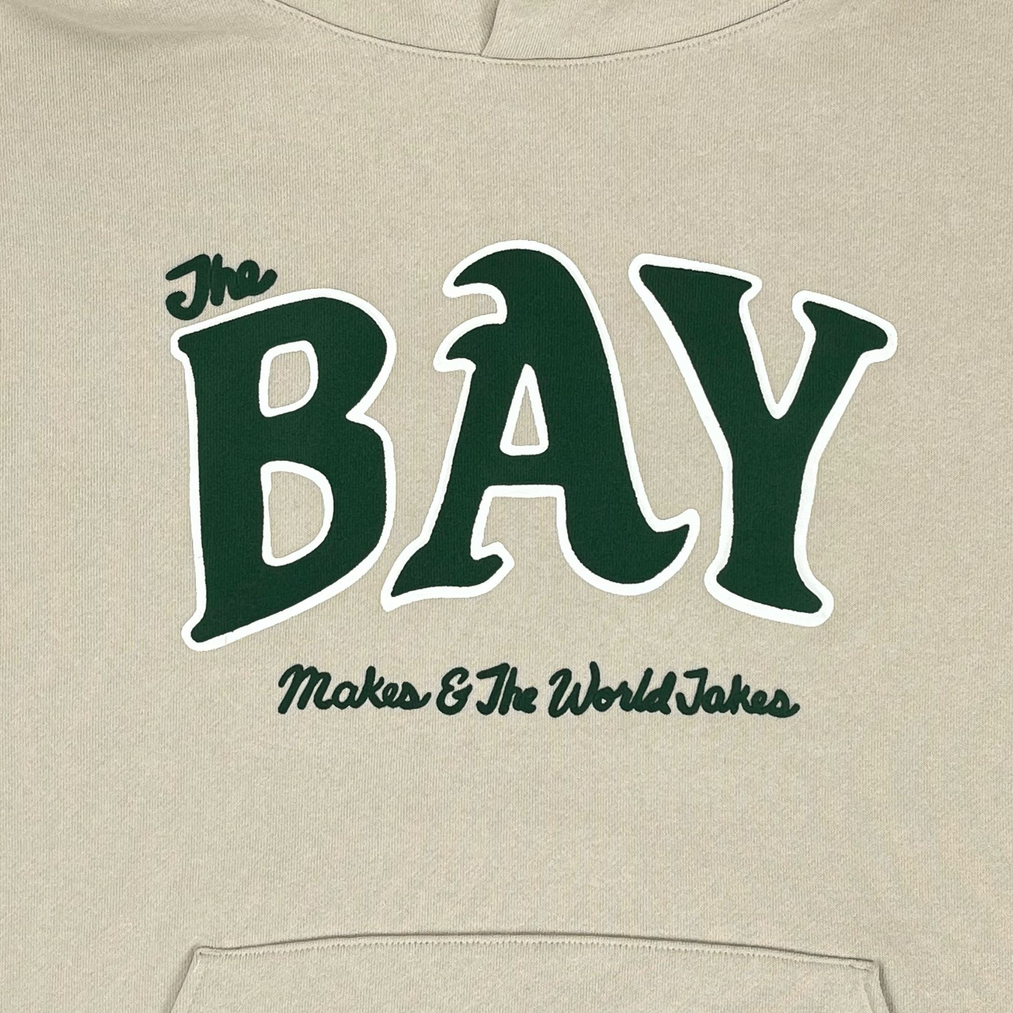 Close-up of a large green The Bay Makes, The World Takes logo on the front chest of an ivory pullover hoodie sweatshirt.