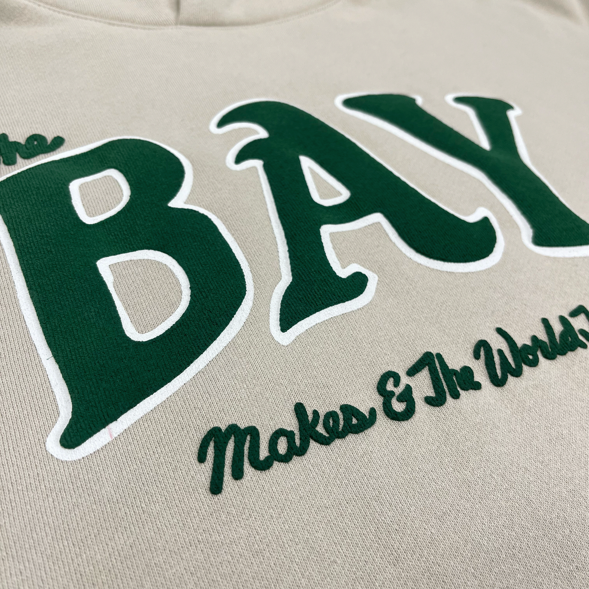 Close-up of green and white The Bay Makes, The World Takes logo on the front chest of an ivory hooded sweatshirt.