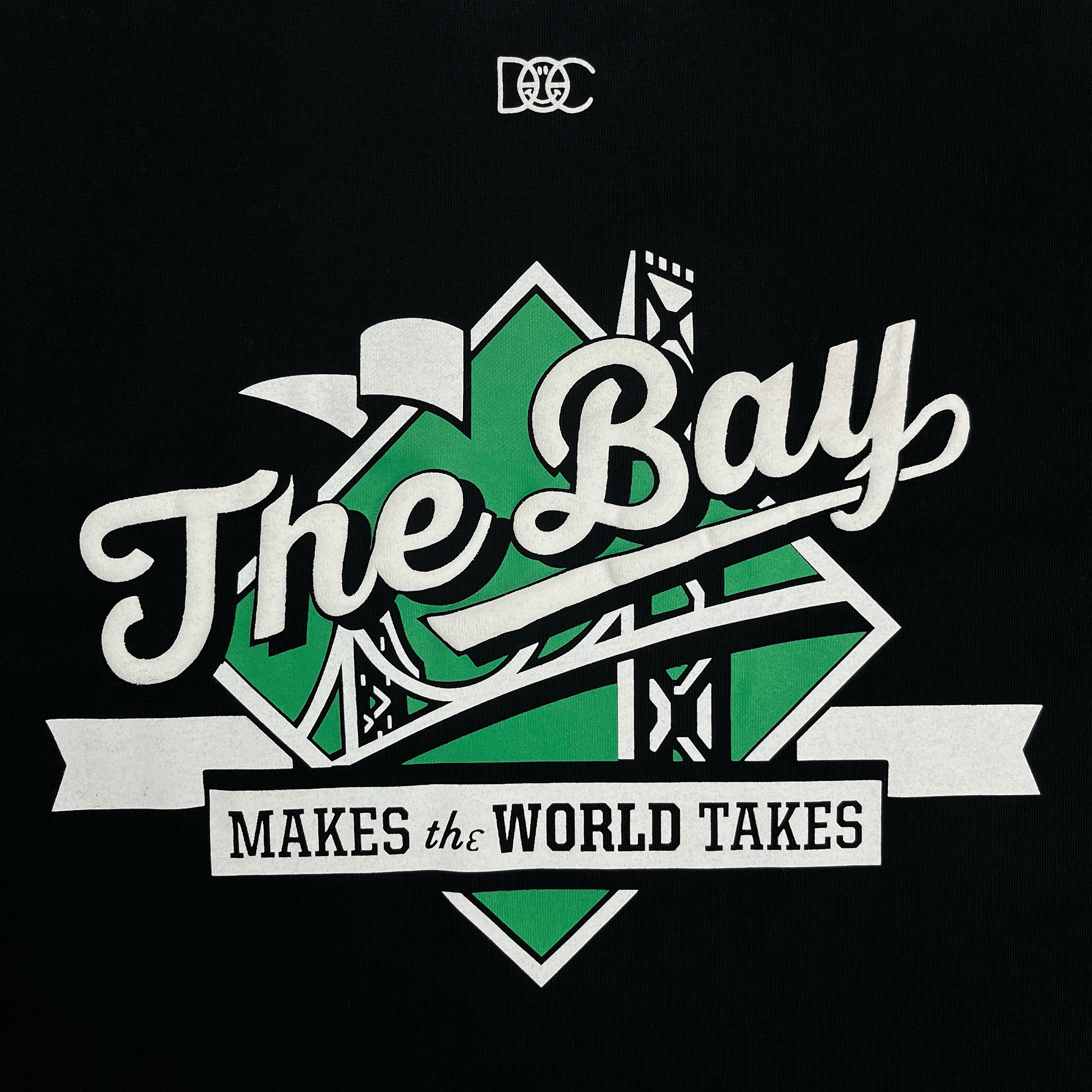 Close-up of The Bay Makes The World Takes green and white logo graphic on a black t-shirt.