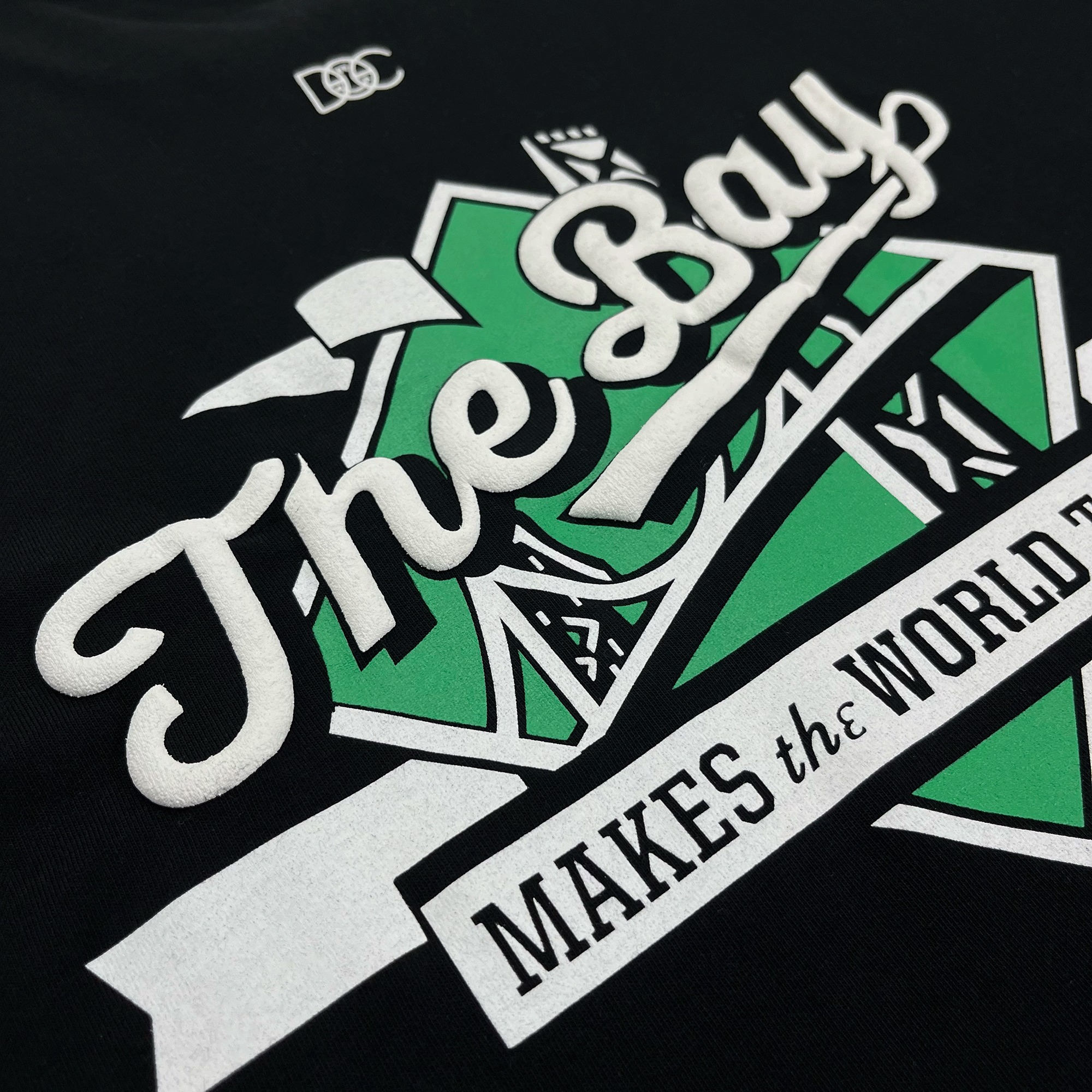 The Bay Tee by DOC from Oaklandish is a classic fit black t-shirt featuring a vibrant design in green and white. The text "The Bay" is prominently displayed in large, cursive font, with "MAKES THE WORLD" written in capital letters below. The design boasts a green geometric shape with abstract elements that celebrate Bay Area culture. Made from 100% cotton, the t-shirt also includes a small "PSC" logo near the hem.
