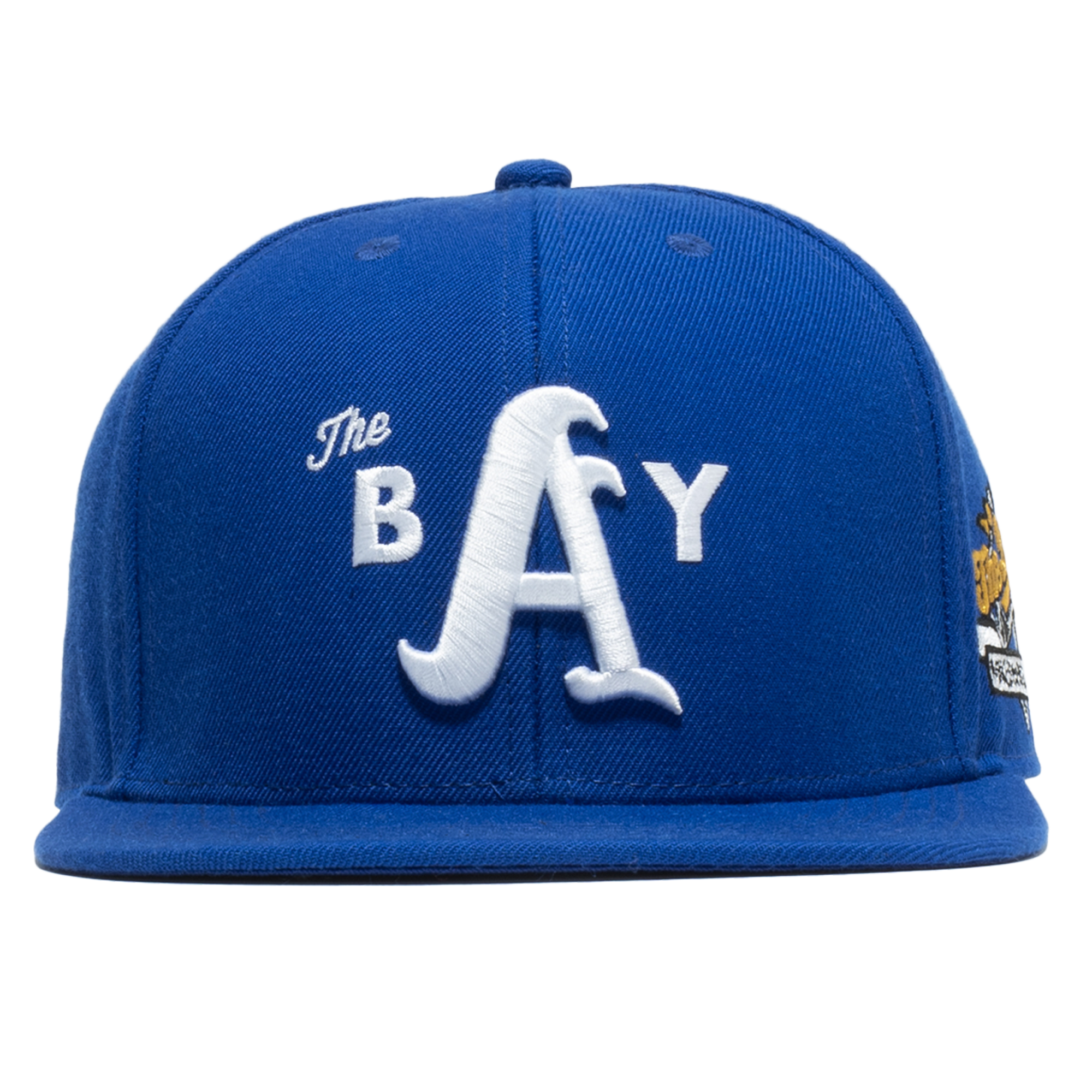 The Bay Snapback by DOC from Oaklandish is a royal blue snapback hat with a flat brim, showcasing white embroidered text and designs on the front. The text reads "The BAY" with the large letter "A" prominently in the center, and an additional multicolored patch is visible on the side of the cap.