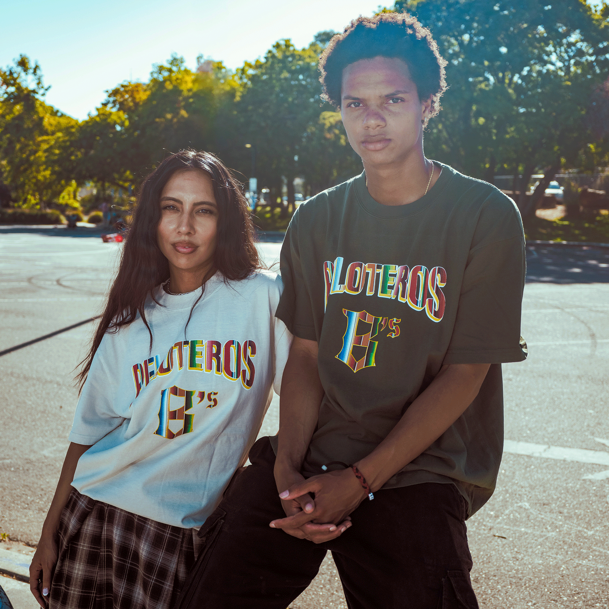Two young adults stand outdoors in sunlight, both wearing Oakland Ballers gear. The woman wears the Oakland Beisbol Heavy Tee with a plaid skirt and leans on the man’s knee. Trees and pavement appear in the background as they face the camera.