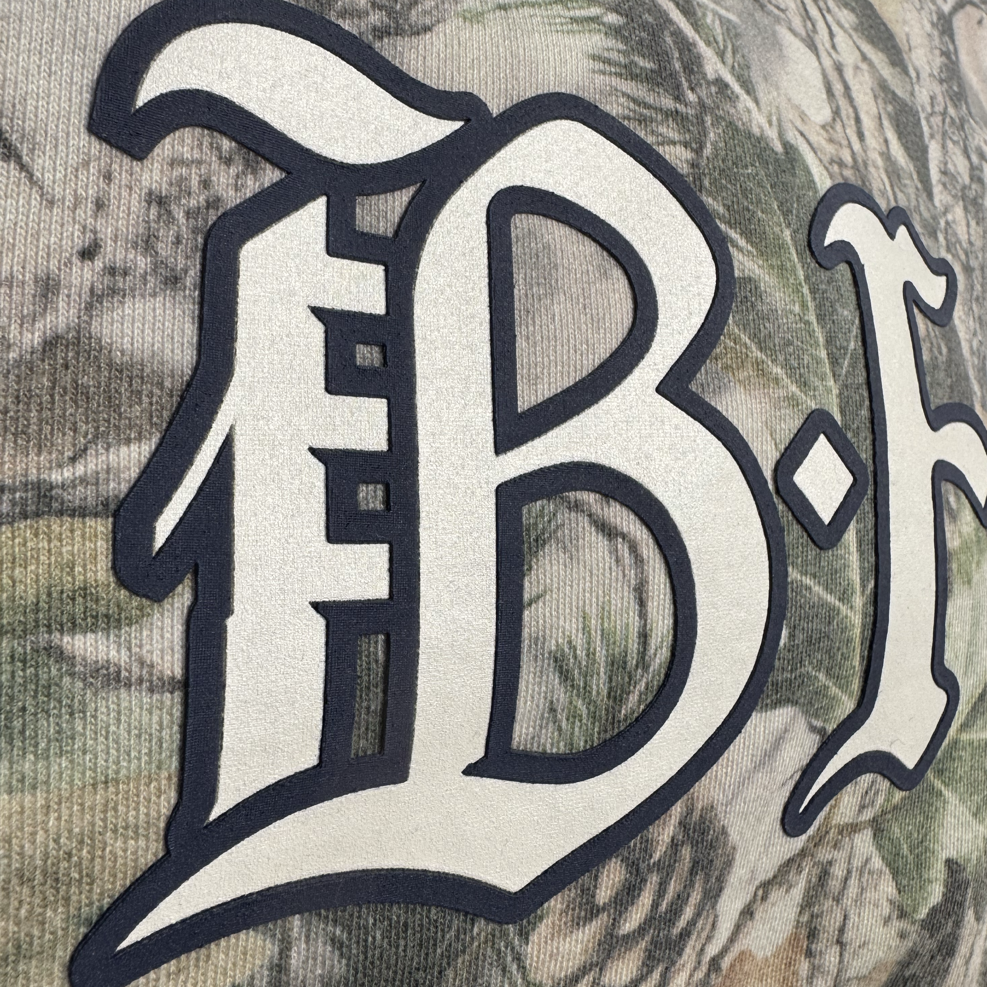 Close-up of a large cream “B” outlined in black on ribbed wood camo fabric with green, brown, and gray shades—detail from the Bay FC Camo Hoodie by Bay FC. Men's pullover hoodie shows partial text on the right.