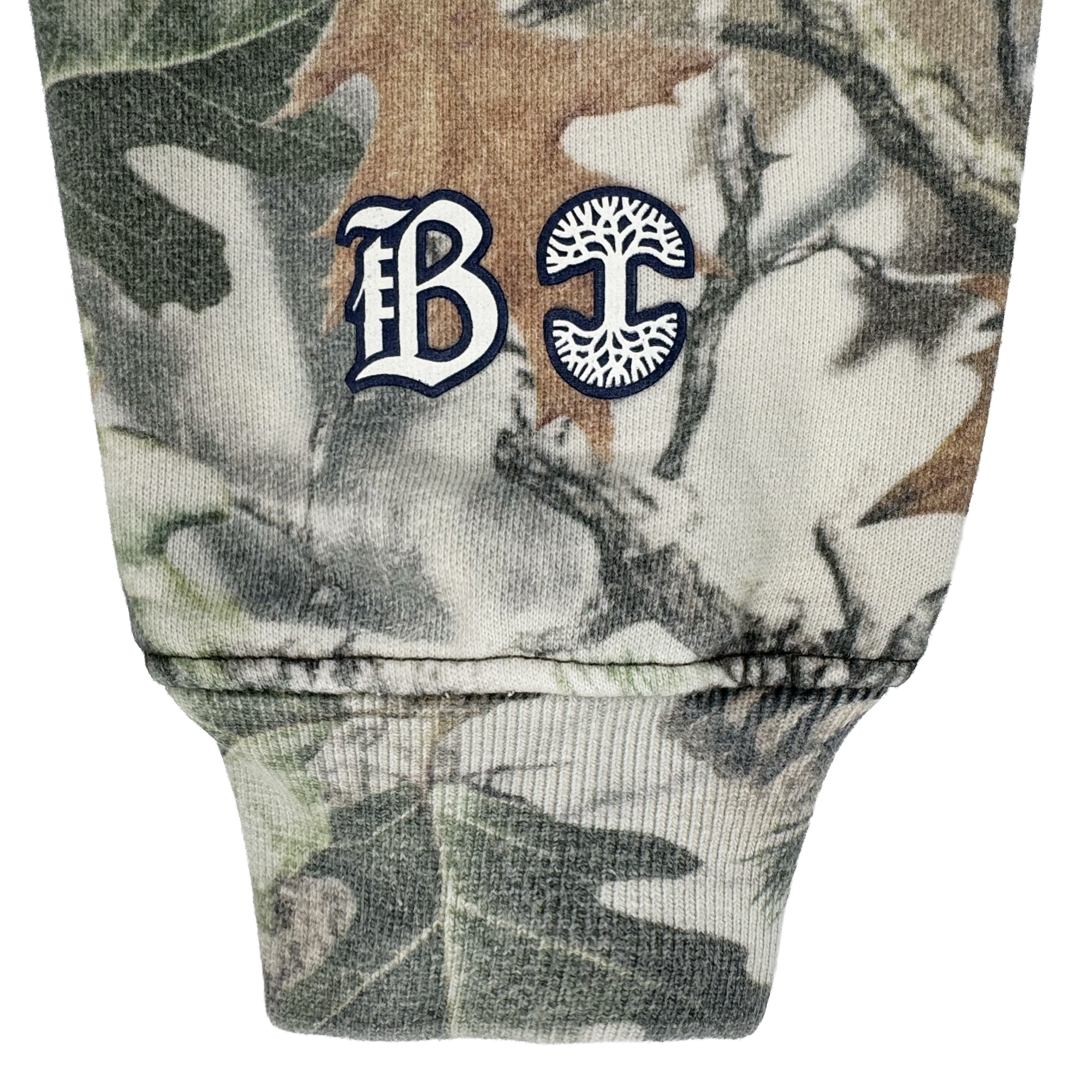 A close-up of the Bay FC Camo Hoodie shows a cuffed sleeve with green, brown, and beige camouflage. Near the seam are white and navy embroidered gothic "B" and a "C" with a tree design, representing Bay FC’s branding.