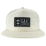 Front view of Ecru nylon snapback cap with embroidered Oaklandish black and white woven patch at center.