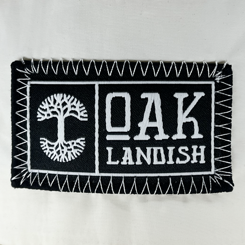 Detailed close up of embroidered Oaklandish black and white woven patch at center of ecru nylon snapback cap.