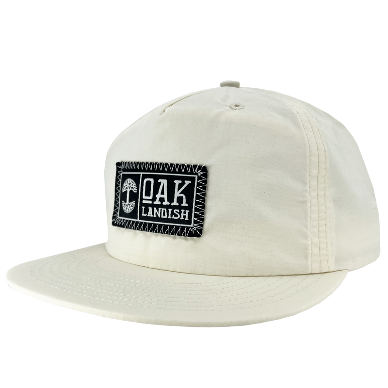Side view of Ecru nylon snapback cap with embroidered Oaklandish black and white woven patch at center.