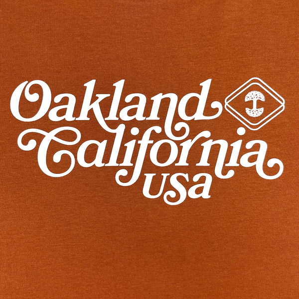 Close-up of Oakland, California, USA graphic in cursive and Oaklandish logo on a red clay-colored t-shirt.