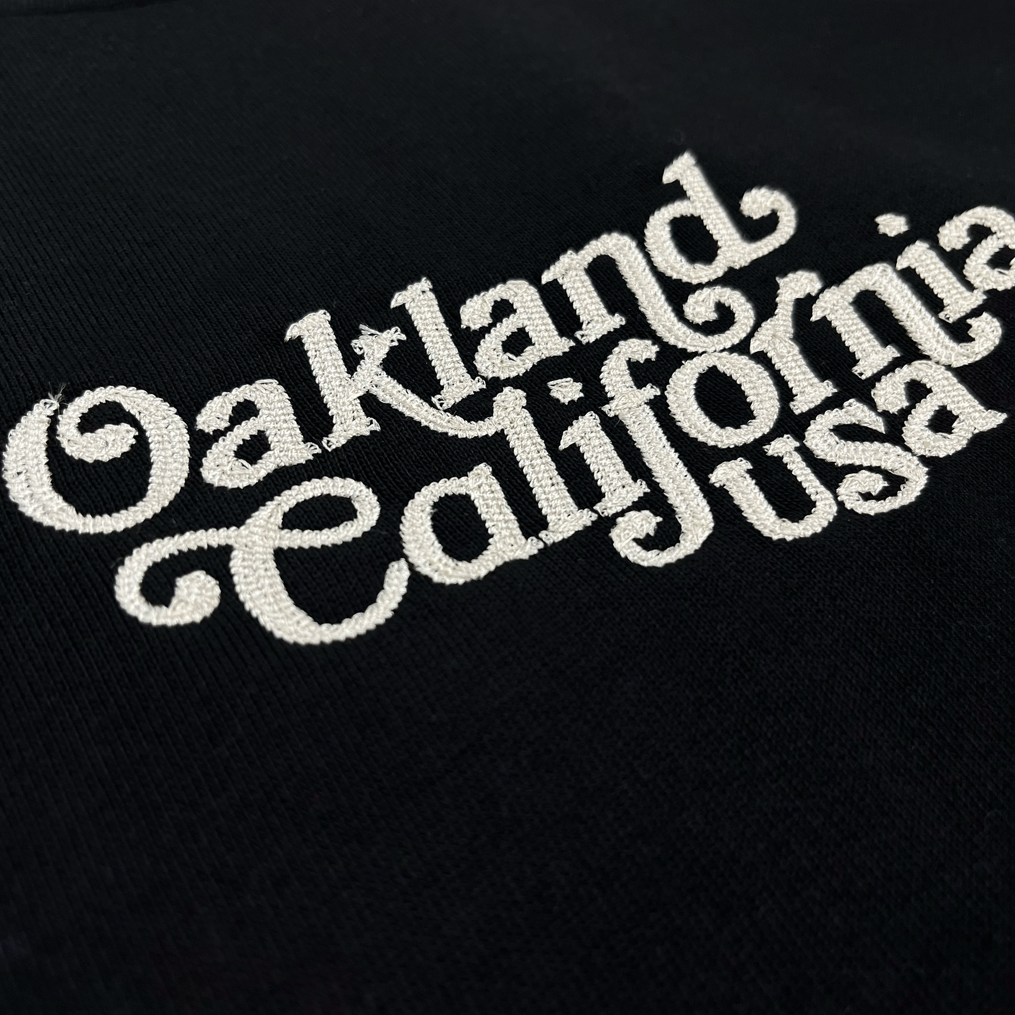 Detailed close-up of white Oakland California USA script embroidered on the chest of a black t-shirt.