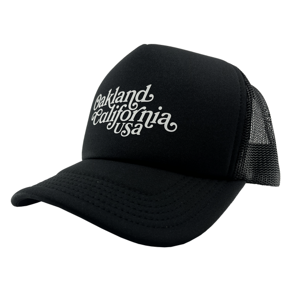 Side view of a black trucker cap with foam front panel, mesh back, adjustable back, and white Oakland, California, USA wordmark on the crown.