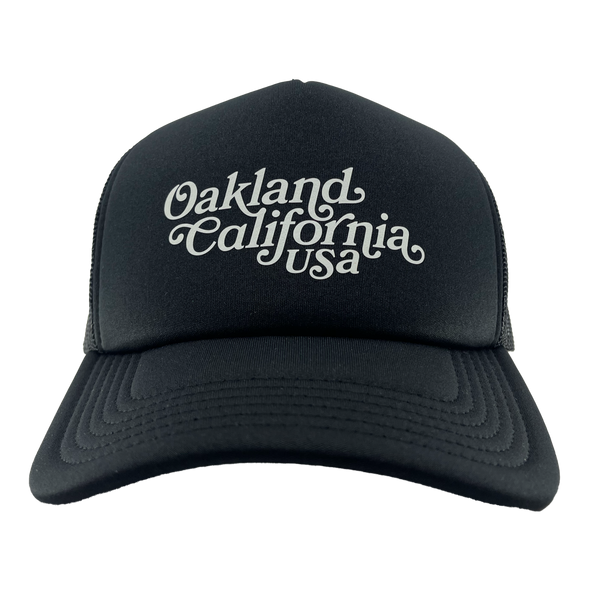 Front view of a black trucker cap with foam front panel, mesh back, adjustable with white Oakland, California, USA wordmark.