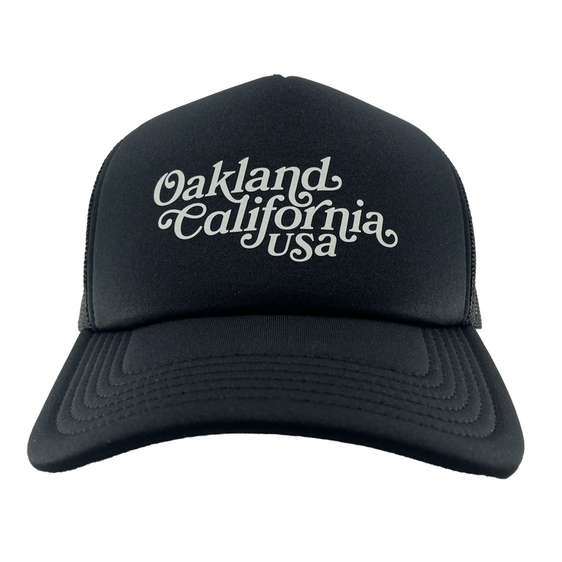Front view of a black trucker cap with foam front panel, mesh back, adjustable with white Oakland, California, USA wordmark.