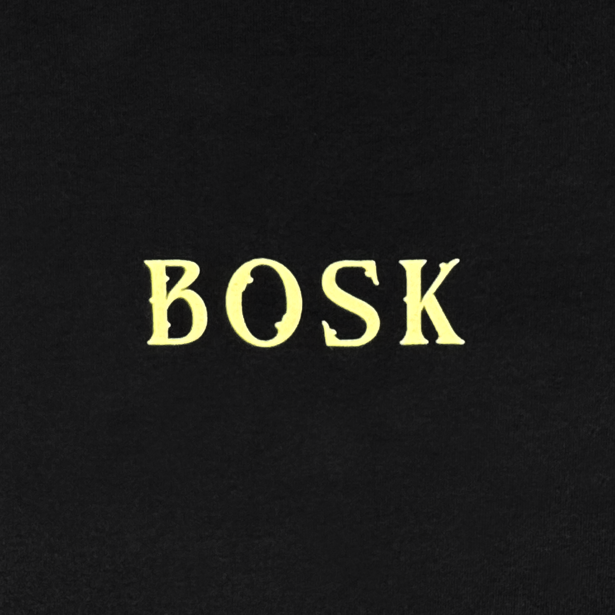 The image showcases "BOSK" in large, pale yellow serif letters on a solid black background, giving a vintage vibe—ideal for the BOSK Allure LS Tee, a 100% cotton long sleeve t-shirt with a classic fit.