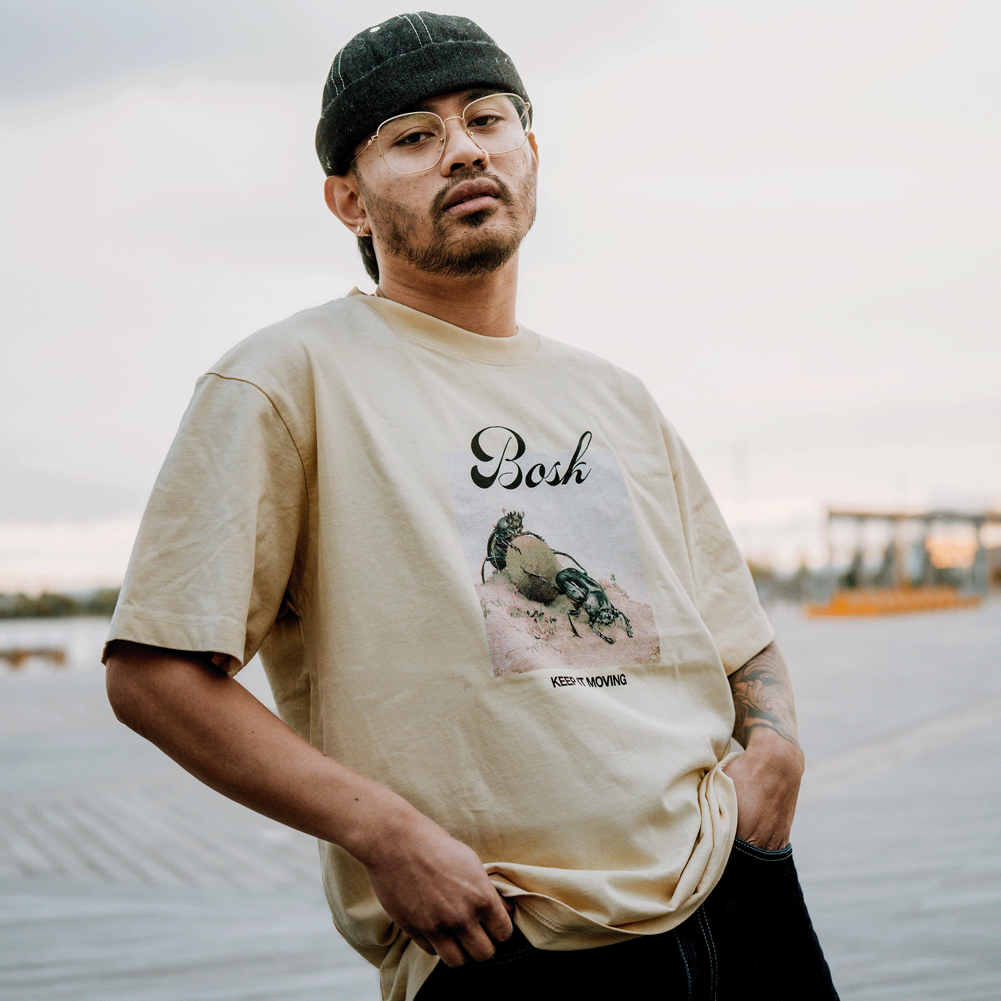 A man in glasses and a black cap stands on a boardwalk, wearing the BOSK Moving Tee—a classic fit, heavy-weight beige T-shirt with a crab graphic and "KEEP MOVING." He looks at the camera, holding his 100% cotton shirt. Background is blurred.