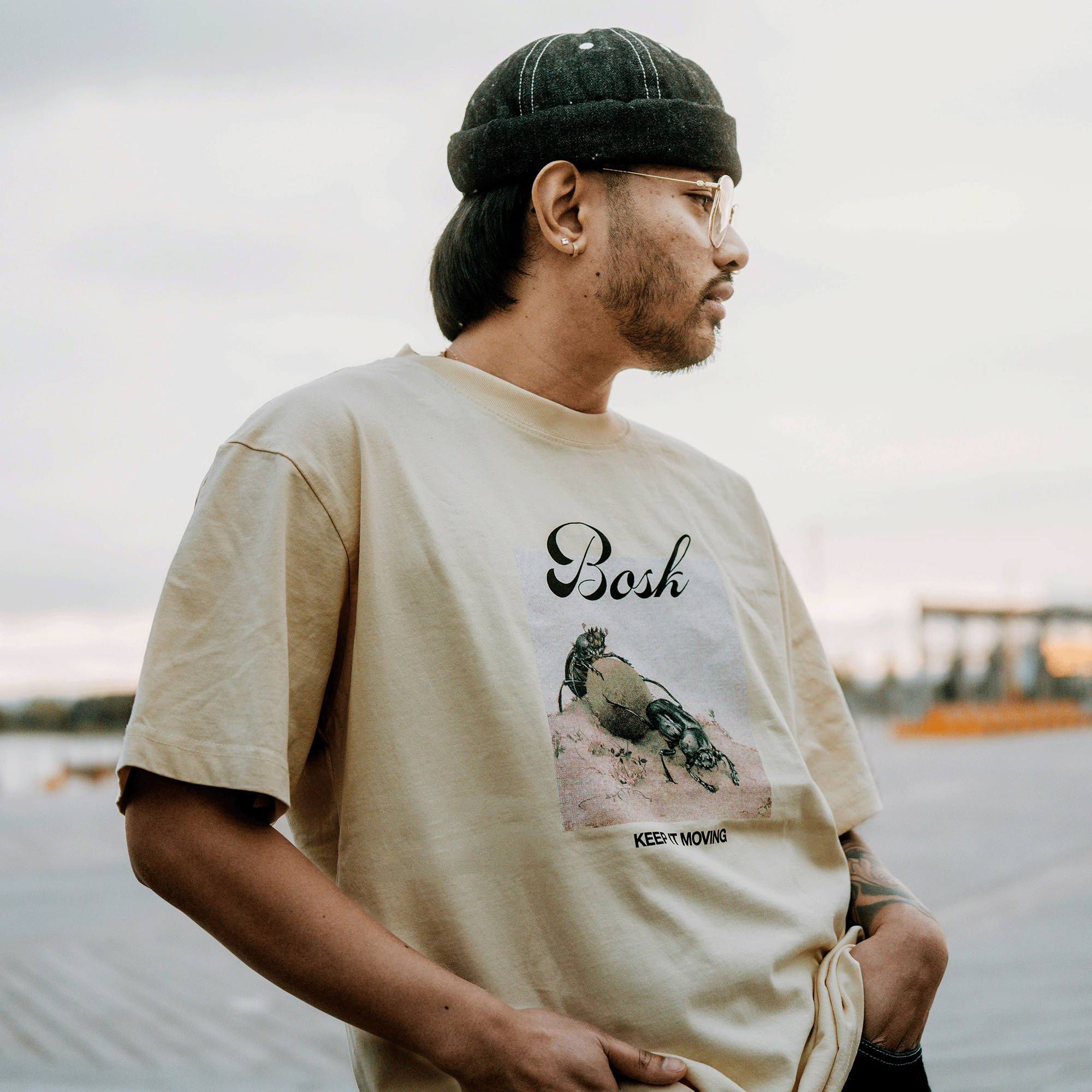 A person with long dark hair, glasses, and a black cap stands outdoors in a classic fit beige BOSK Moving Tee made from heavyweight cotton. The tee displays "BOSK," "Keep Moving," and a turtle graphic; the cloudy sky is blurred behind them.