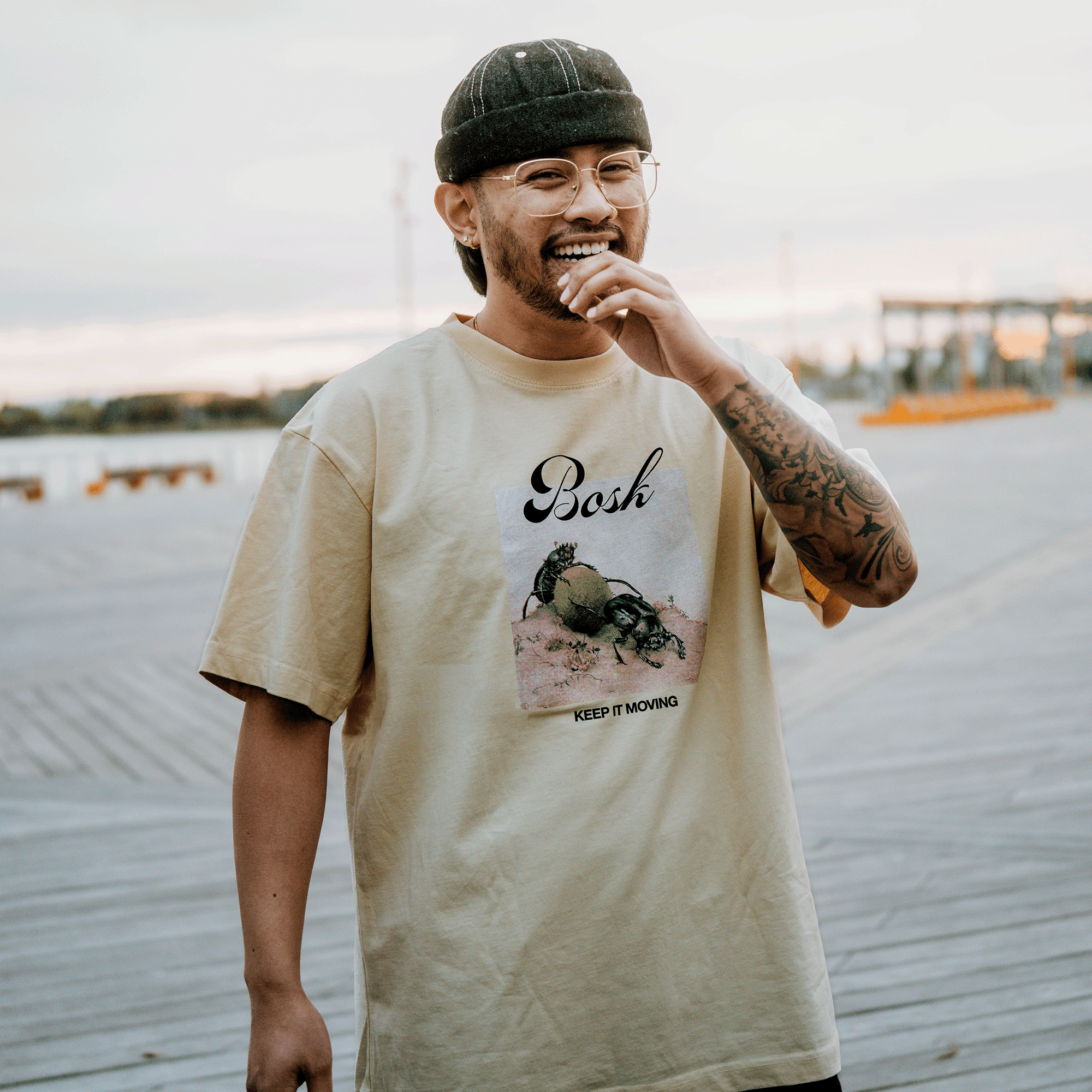 A smiling person in glasses and a dark beanie stands on a boardwalk at sunset, wearing the BOSK Moving Tee— a classic fit heavy-weight cotton T-shirt with a crab graphic and “Bosh” and “Keep It Moving” text. Tattoos show on their right forearm.