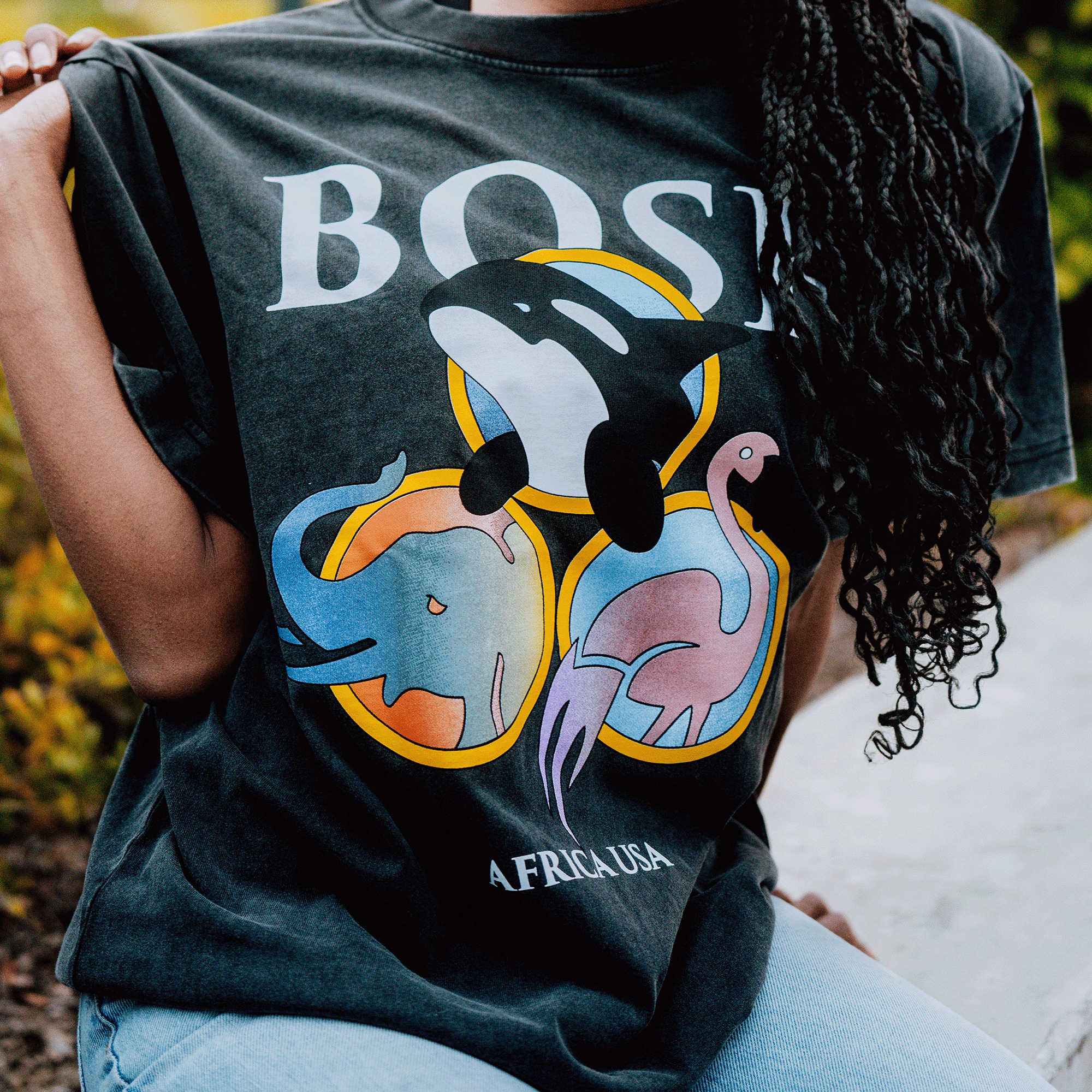 A person wears the BOSK Bosk World Tee, a classic fit 100% cotton shirt with colorful orca, elephant, and flamingo illustrations in yellow circles. “BOSS” is printed above “AFRICA USA.” The person’s face is not shown.