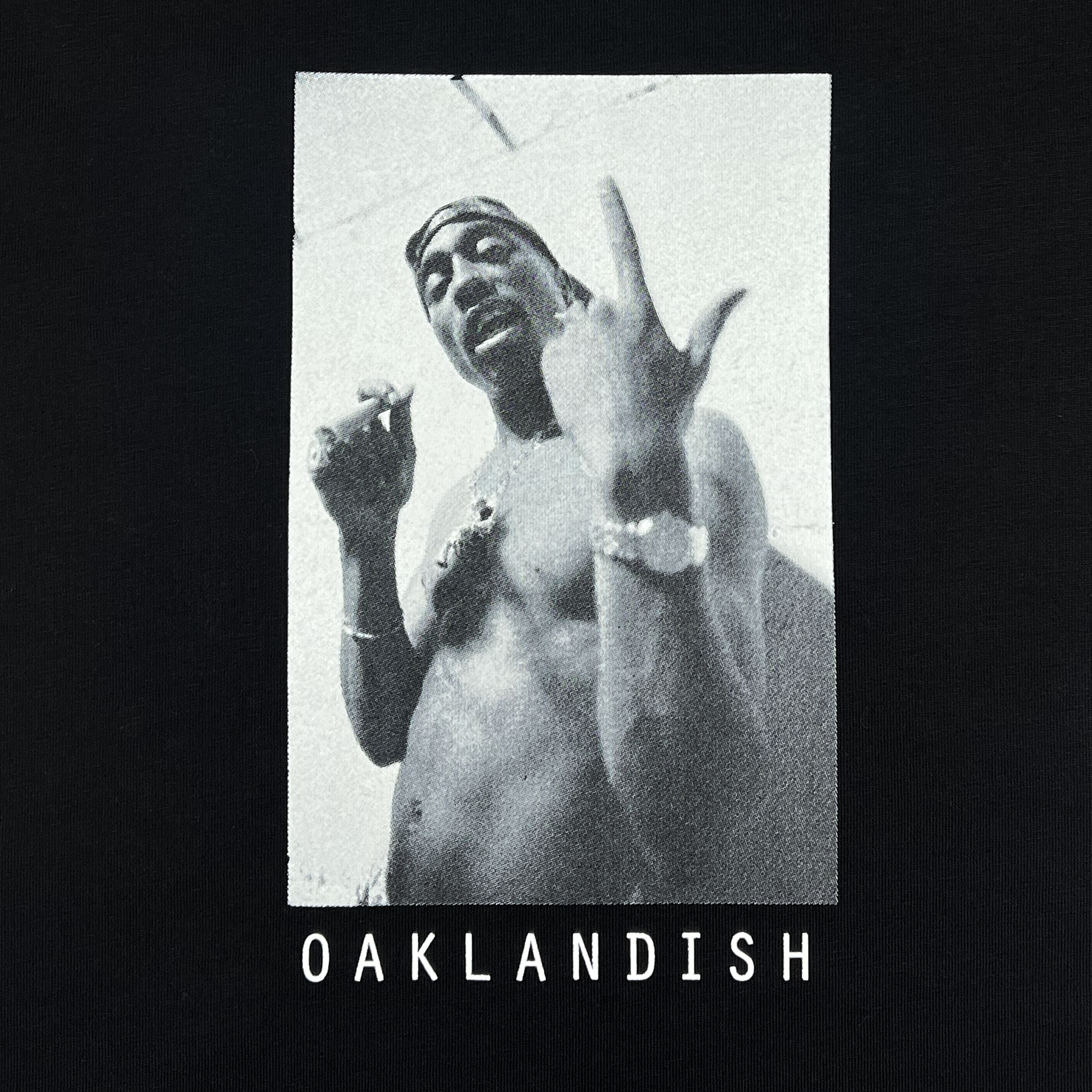 The image captures a shirtless man in black and white, evoking the style of Tupac Shakur with sunglasses, a bandana, multiple necklaces, and a watch, while making a hand gesture. Boldly printed below is "OAKLANDISH," embodying the spirit of their Brighter Days Tee from the hip-hop collection.